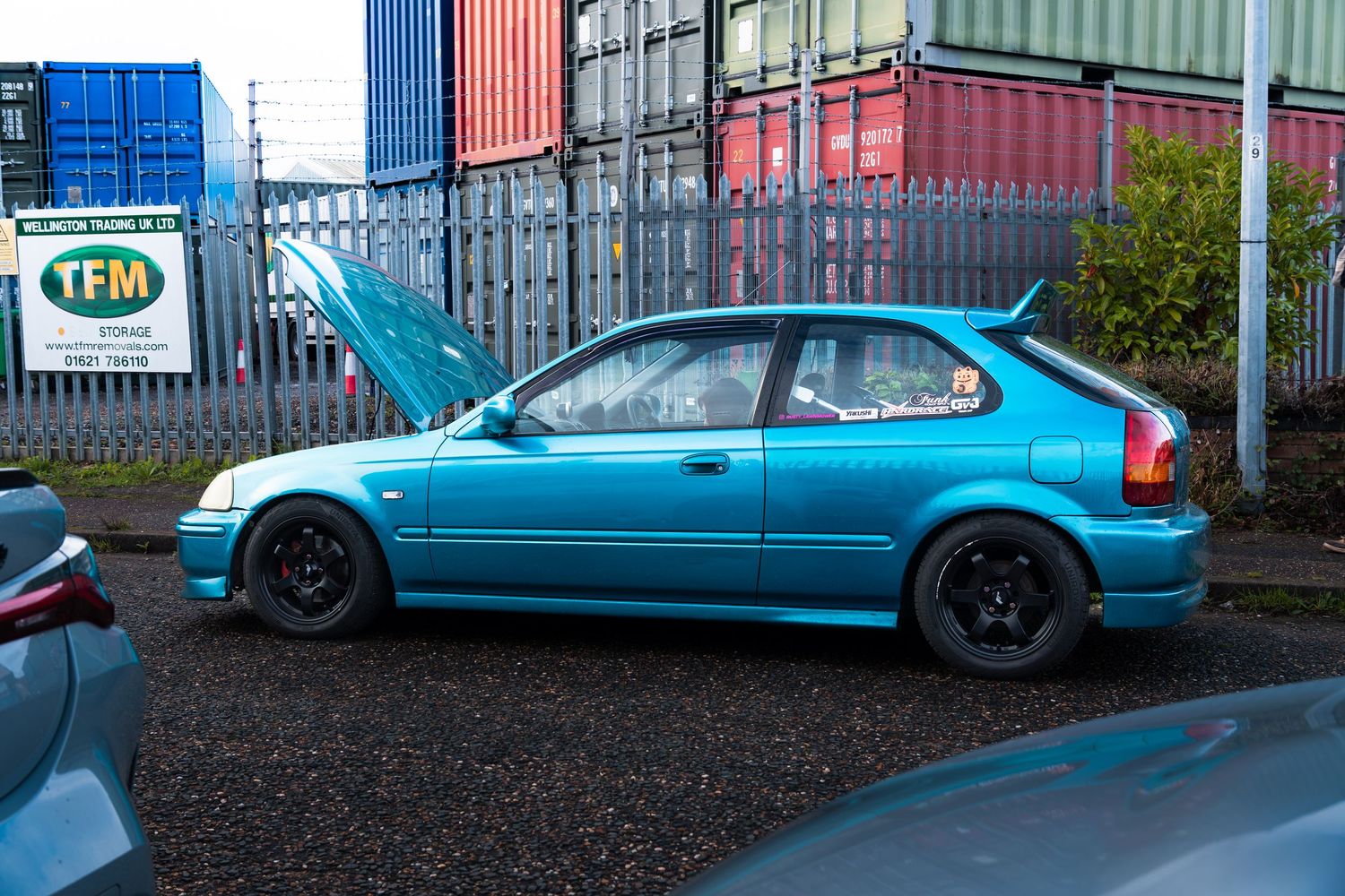 A teal Honda Civic hatchback with black alloys parked with hood open near shipping containers.