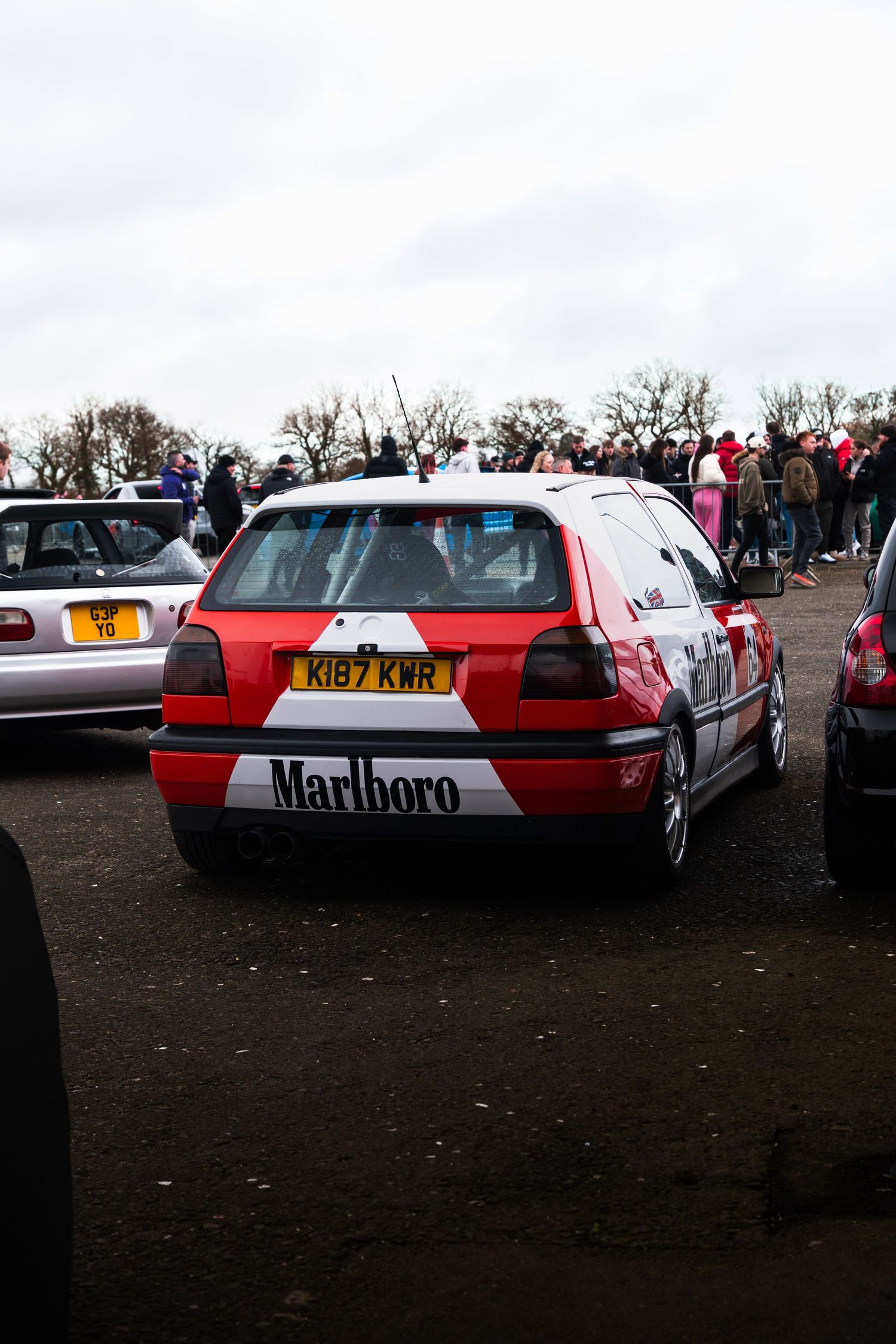 A red and white Marlboro-liveried Volkswagen Golf hatchback parked at an outdoor car meet event.