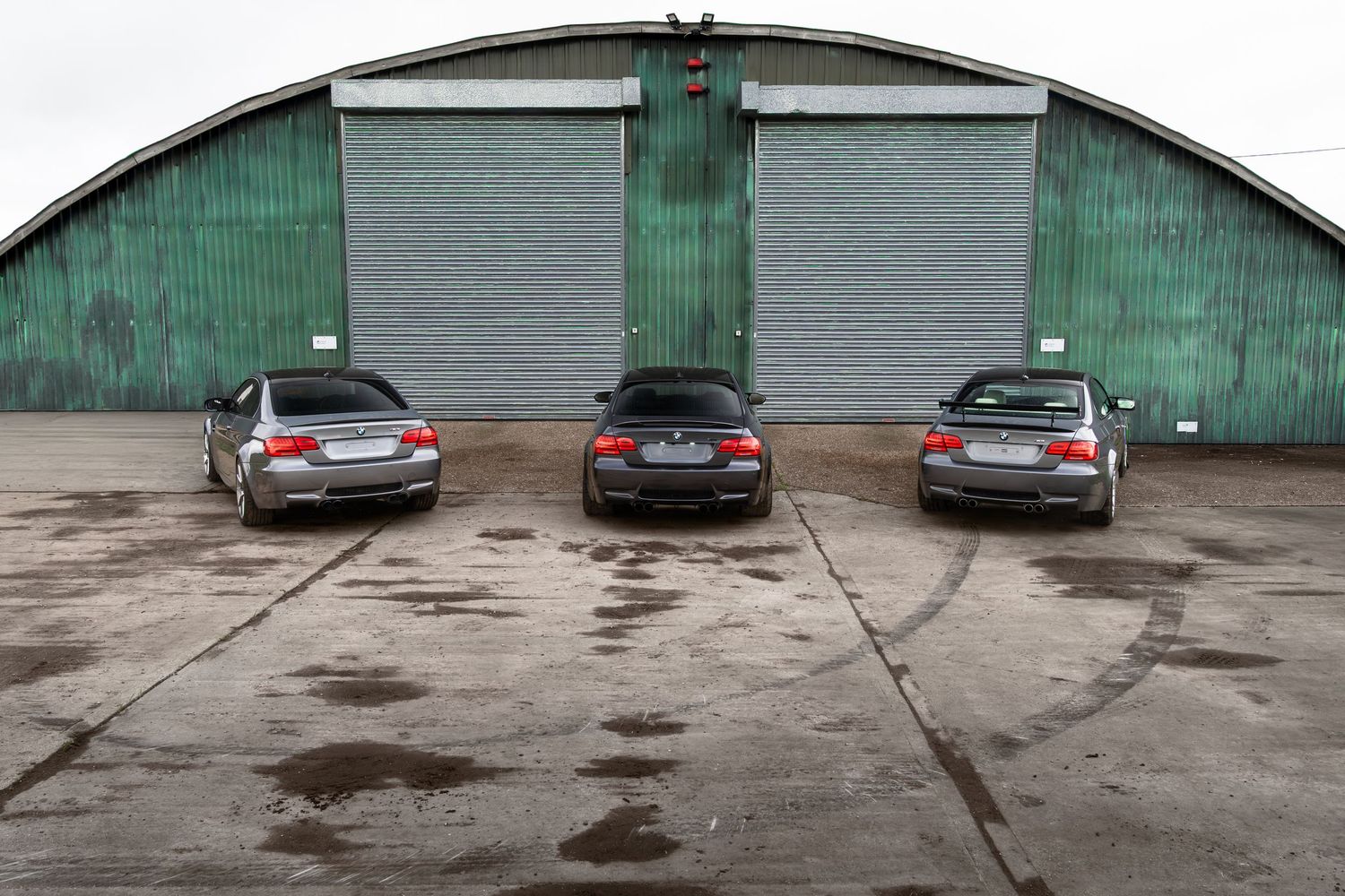 Three BMW M3 coupes seen from behind parked on wet concrete before a green arched hangar.