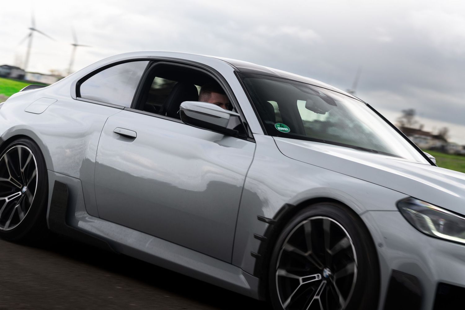 Side profile of a modified white BMW M2 coupe in motion on a track with wide body kit and dark alloy wheels.