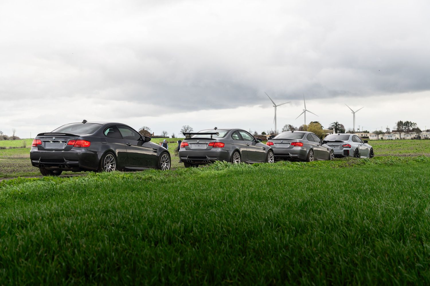 Group of BMW M3 coupes parked in green field with wind turbines in background.