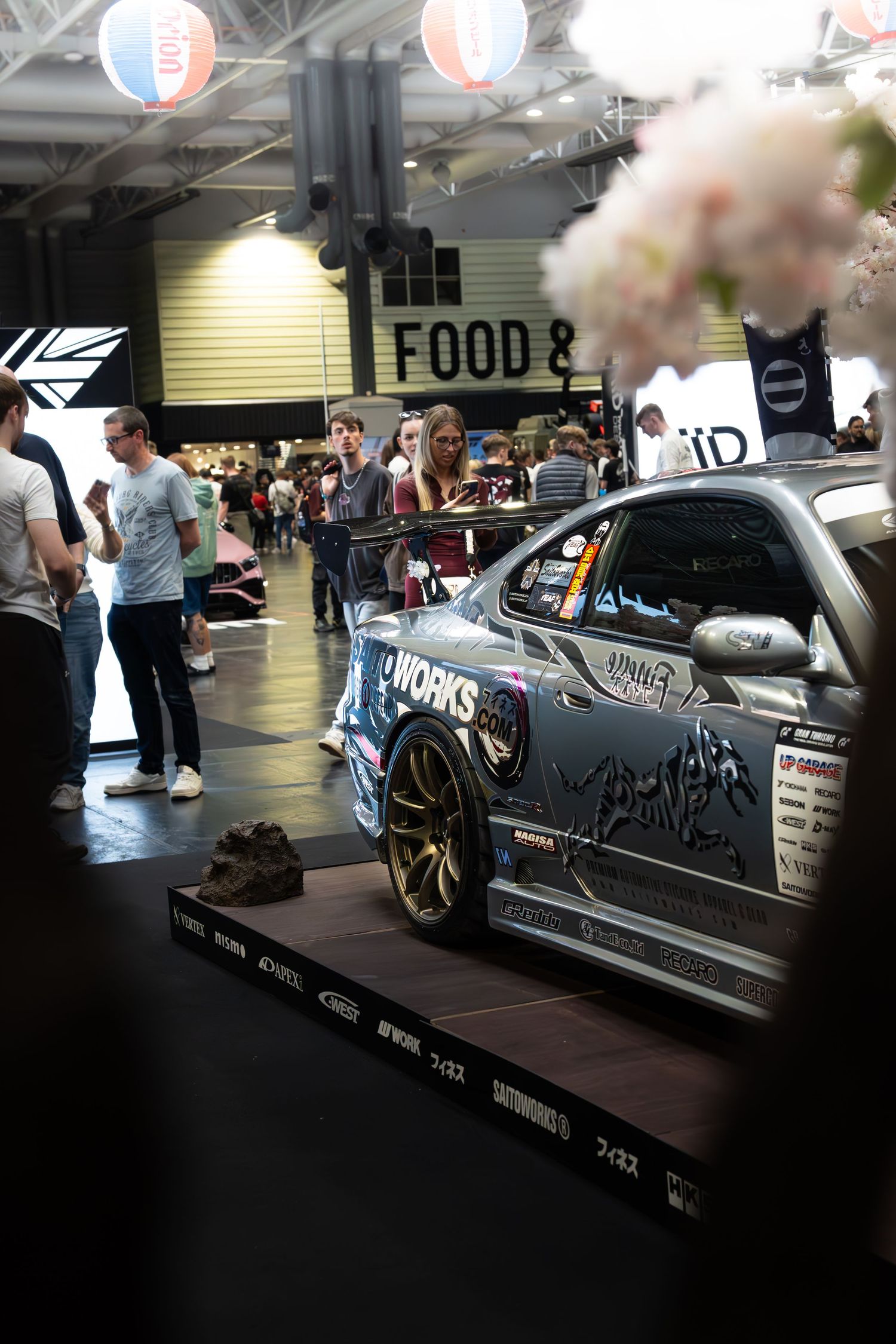 Saitoworks silver drift car on display at indoor car show with spectators and food area in background.