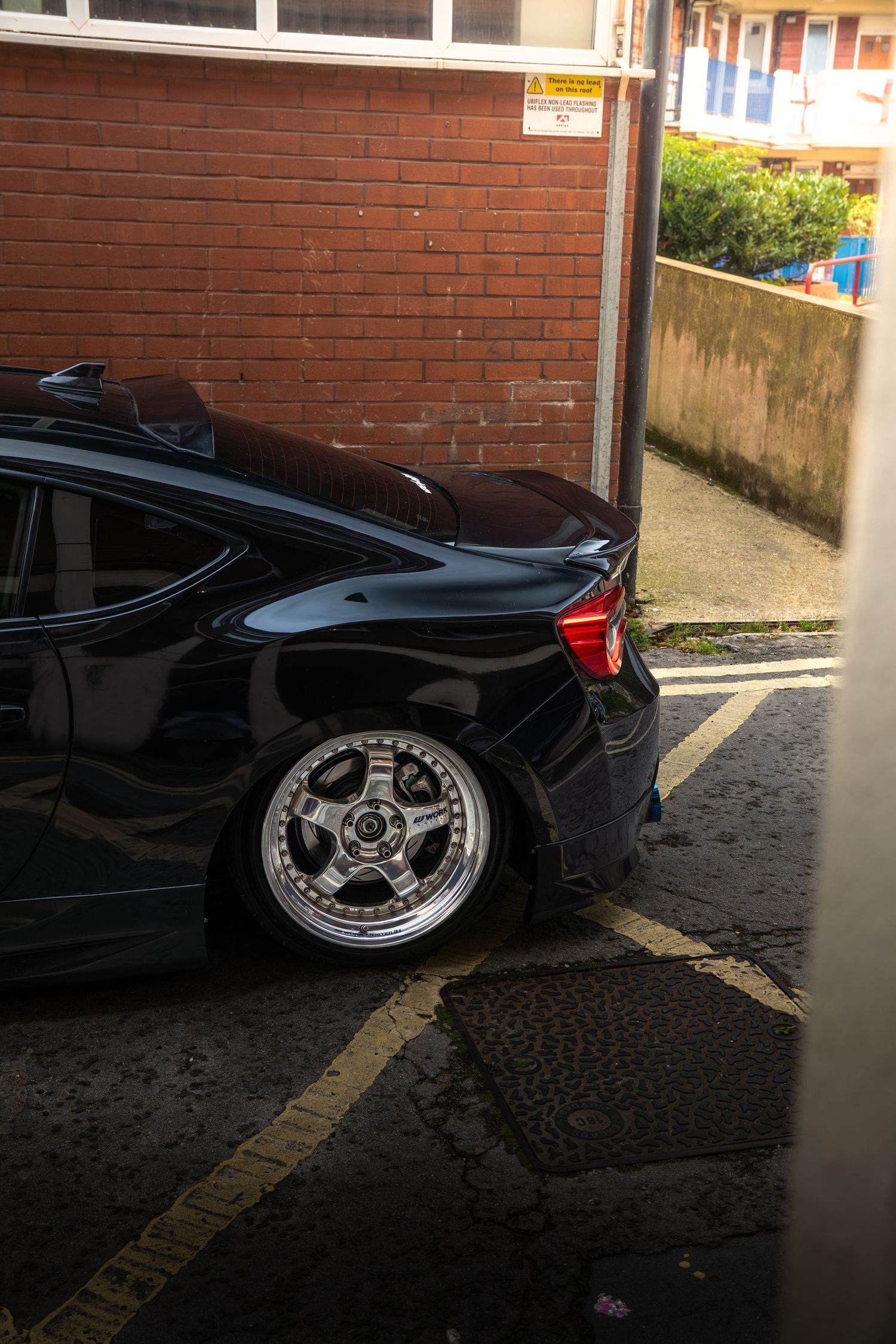 Rear view of a lowered black car with deep-dish chrome wheels on a brick-walled parking area.