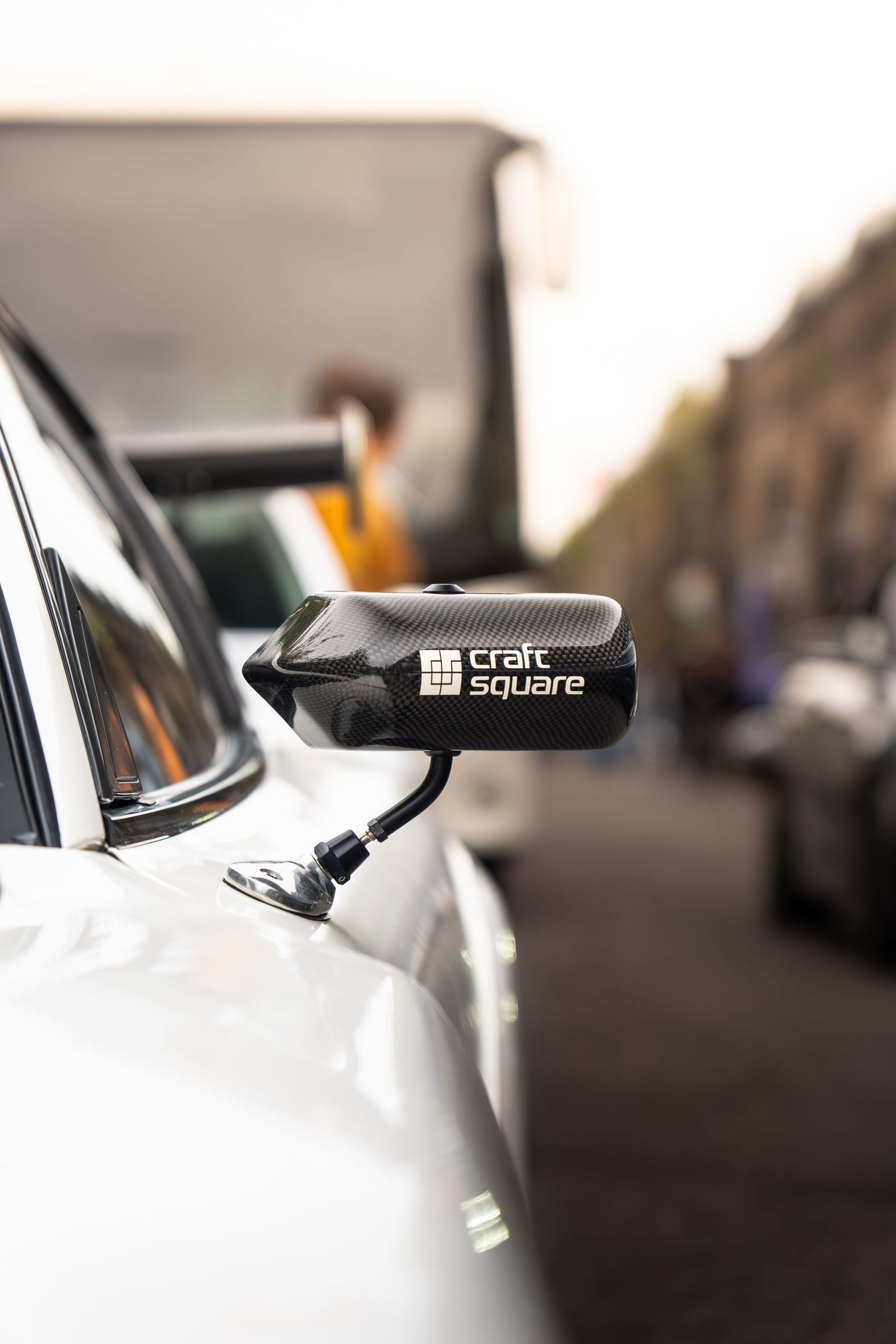 A carbon fiber Craft Square side mirror mounted on a white car in an urban street setting.
