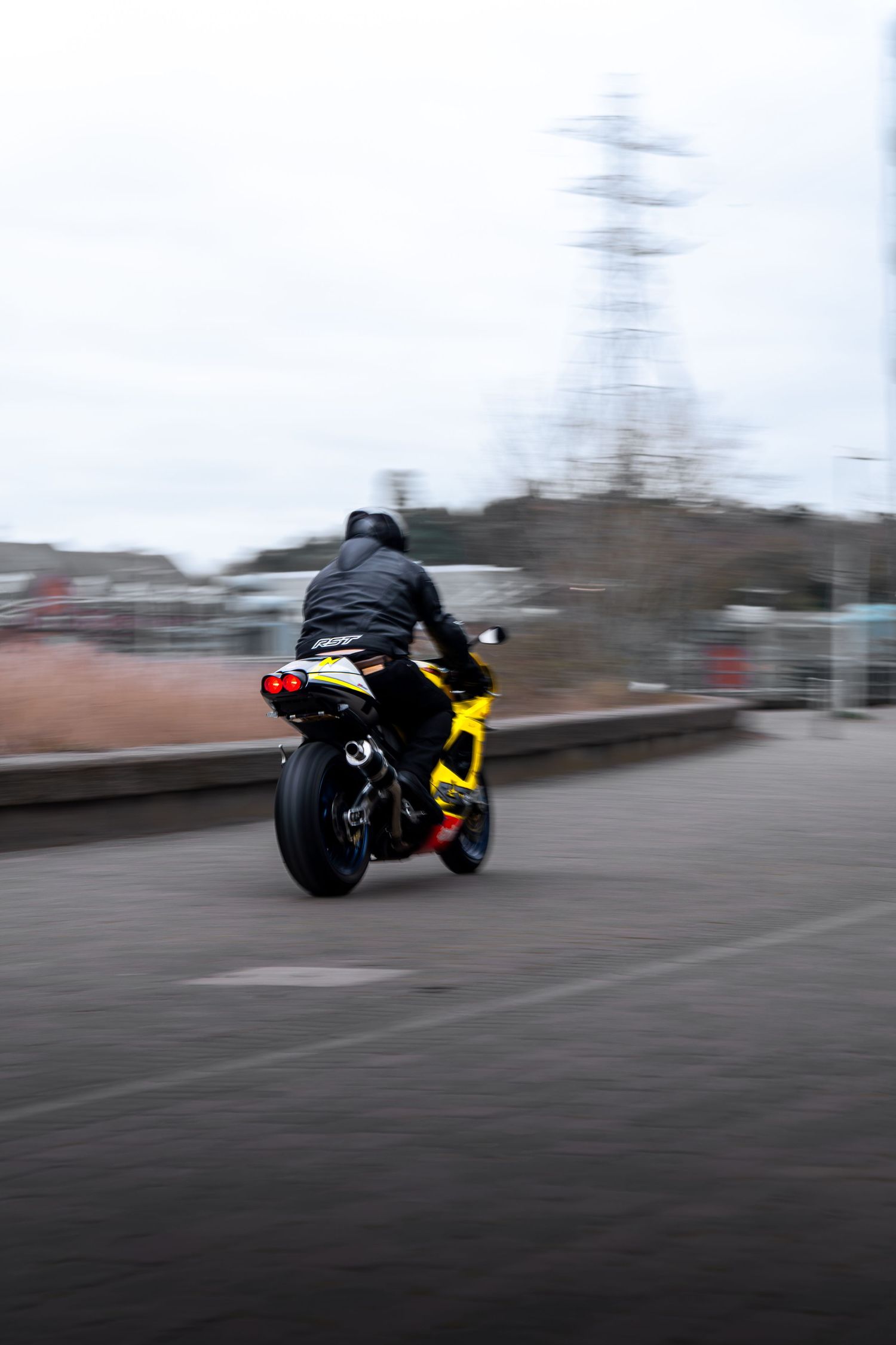 Rear view of a yellow Aprilia superbike accelerating away with a power pylon in background.