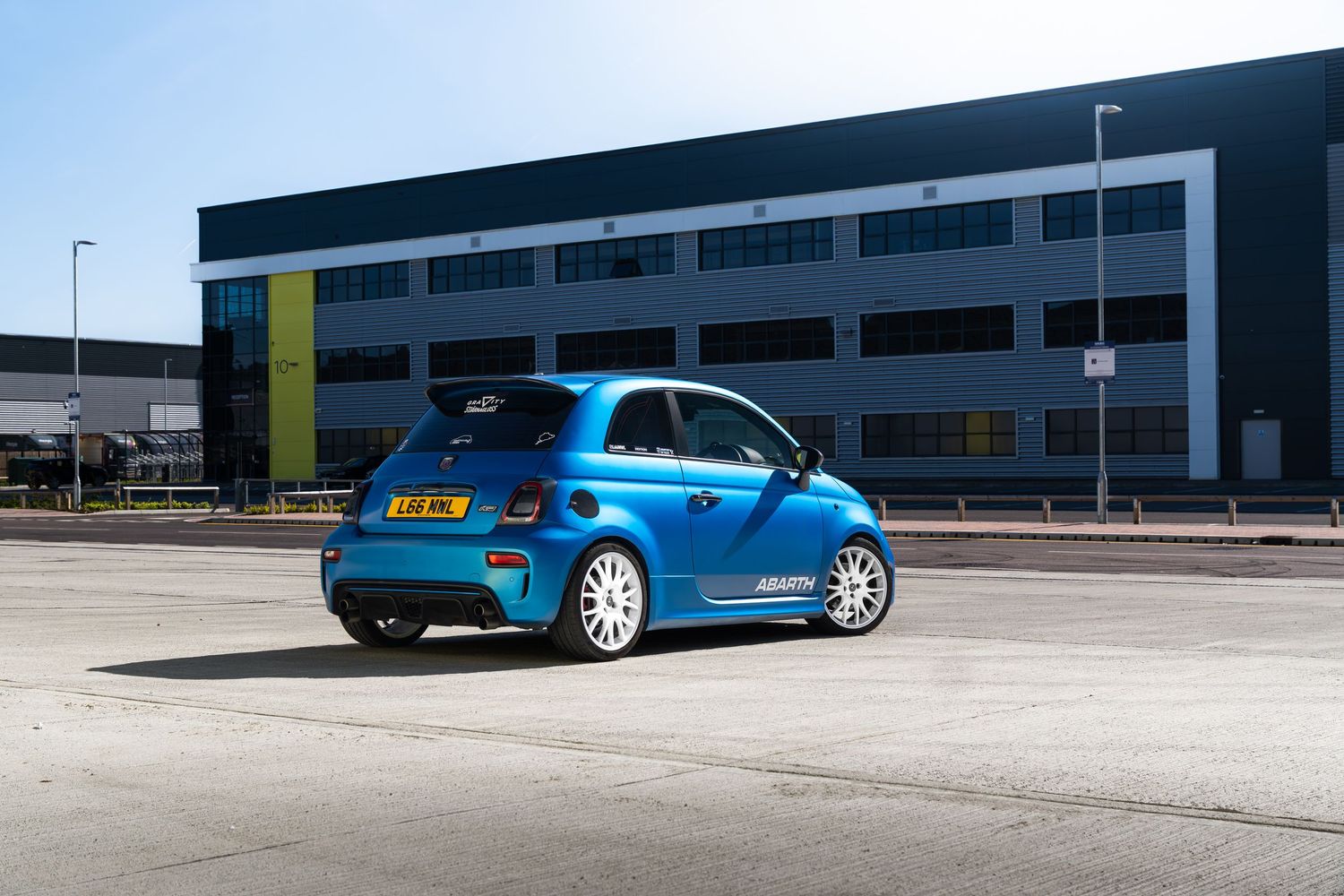 Blue Abarth 595 hatchback parked outside an industrial building, rear three-quarter view on a sunny day.