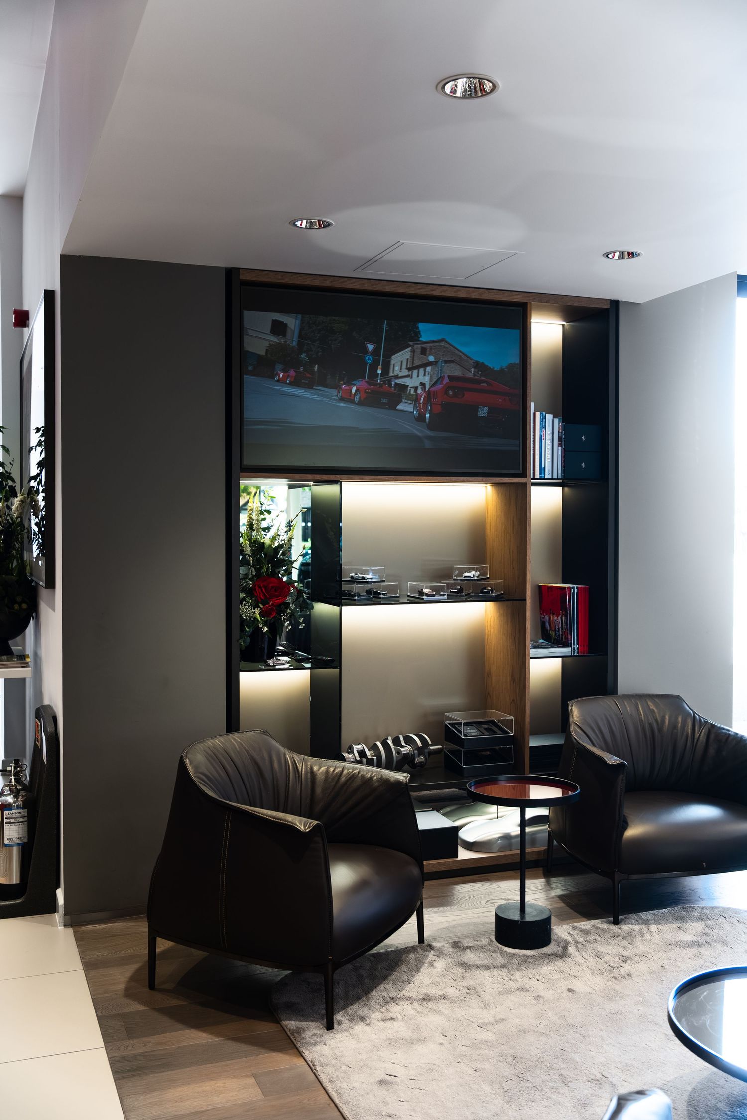 Luxury car dealership lounge with black leather chairs, illuminated shelving, and a large display screen showing red cars.