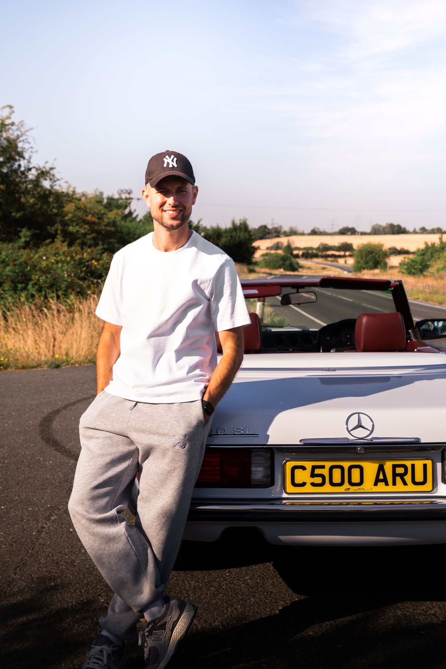 Man in NY cap and white tee leaning on a silver Mercedes convertible with UK plate C500 ARU on a sunny day.