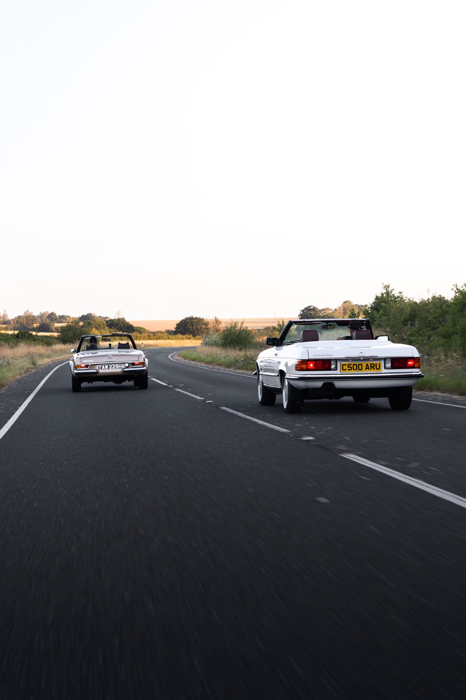 Two white convertible cars driving on a rural road surrounded by fields and trees at dusk.