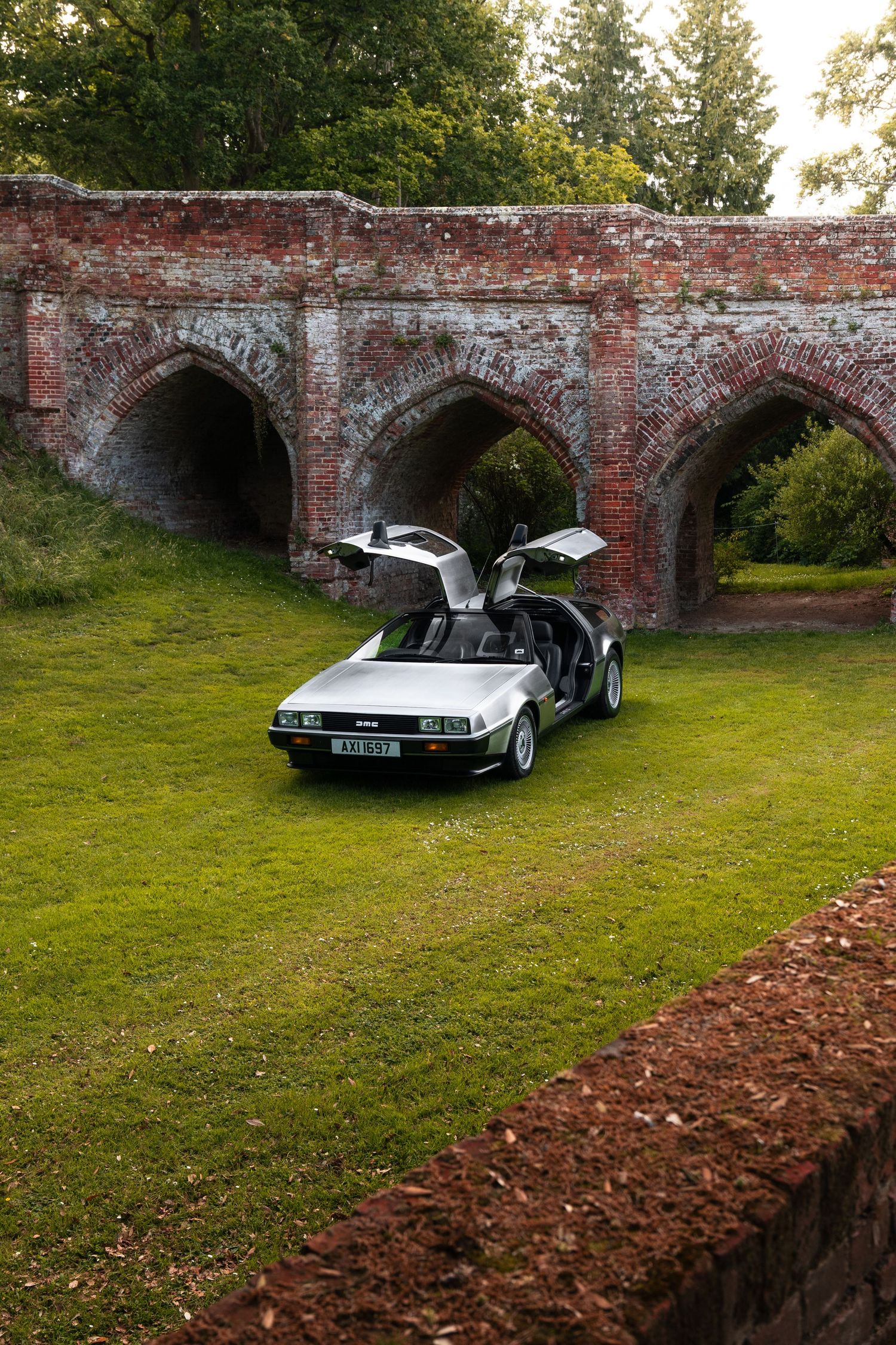 A silver DeLorean DMC-12 with gull-wing doors open, parked on green grass before a historic red brick arched bridge.