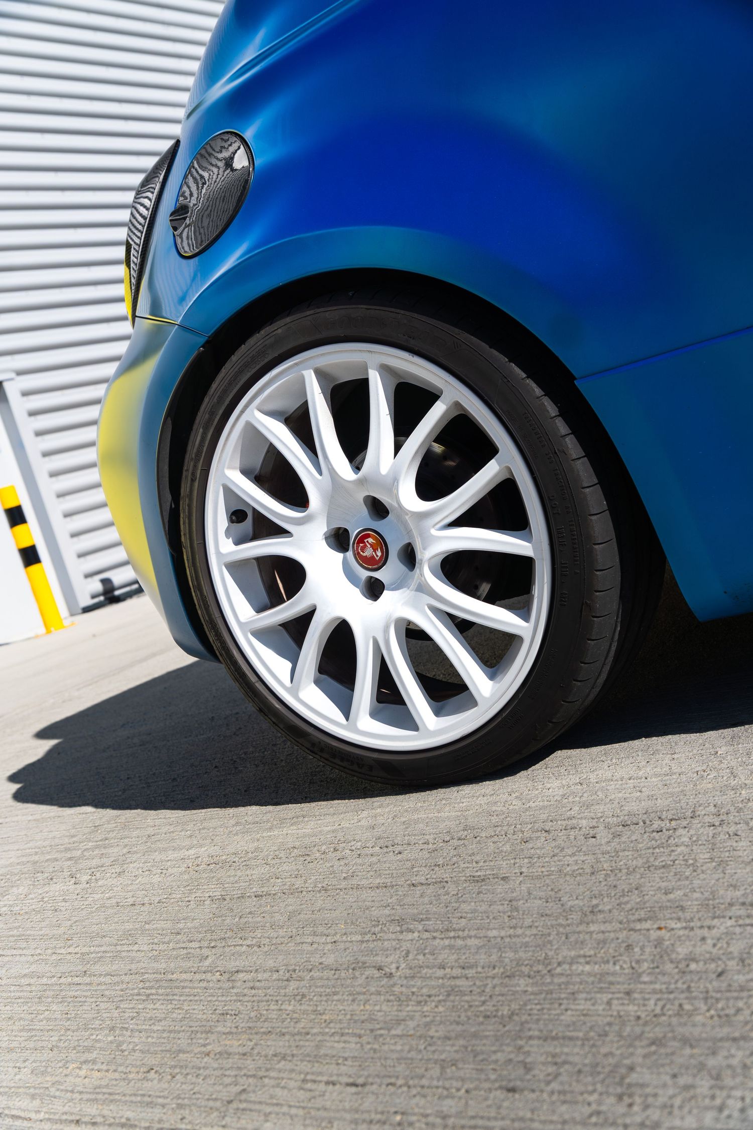 White multi-spoke alloy wheel on a blue Abarth 595 parked on concrete near a yellow garage door.