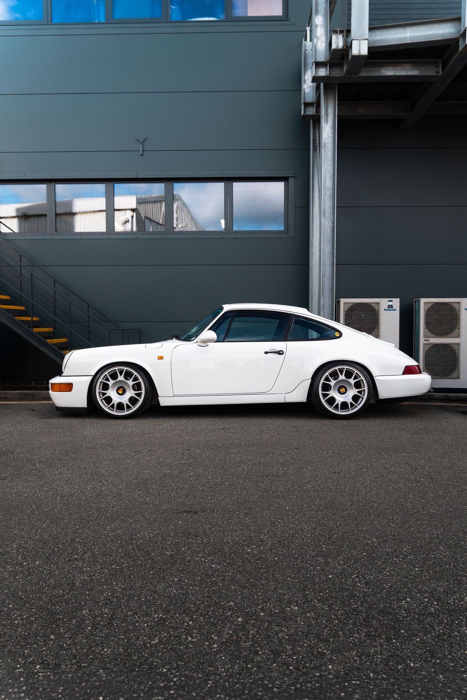 White Porsche 911 964 parked beside industrial building, side profile view.