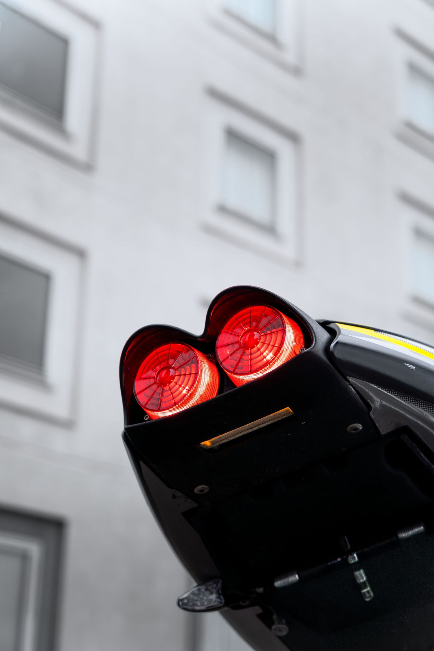 Dual red tail lights glowing on a black sport motorcycle against a grey building backdrop.
