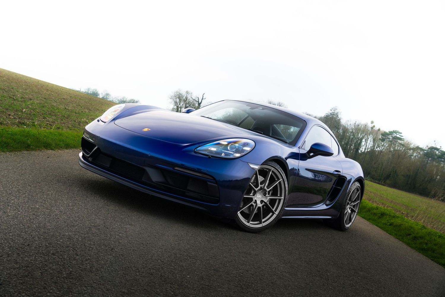 Dynamic low-angle view of blue Porsche 718 Cayman on a curved country road.
