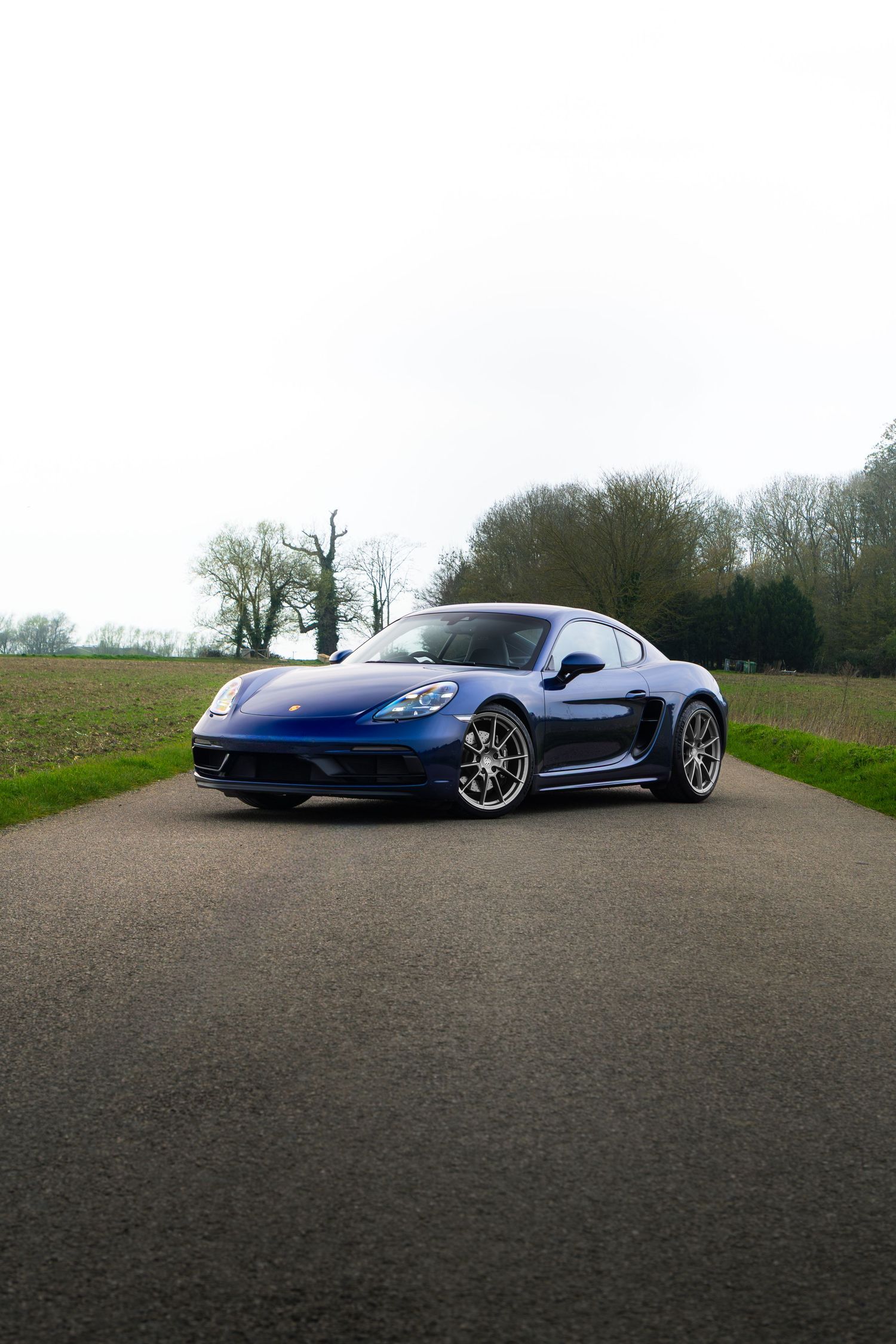 Blue Porsche 718 Cayman angled on a rural driveway with bare trees and fields in background.