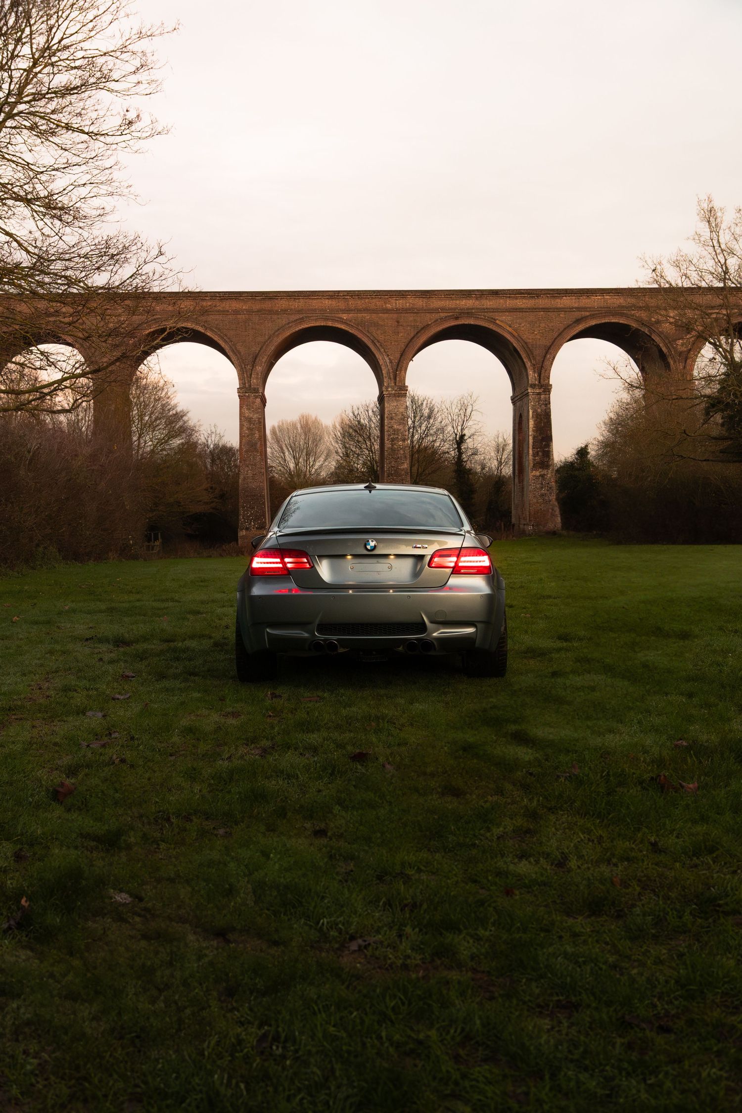 BMW M3 coupe rear view centred before large brick arched viaduct on open grass.