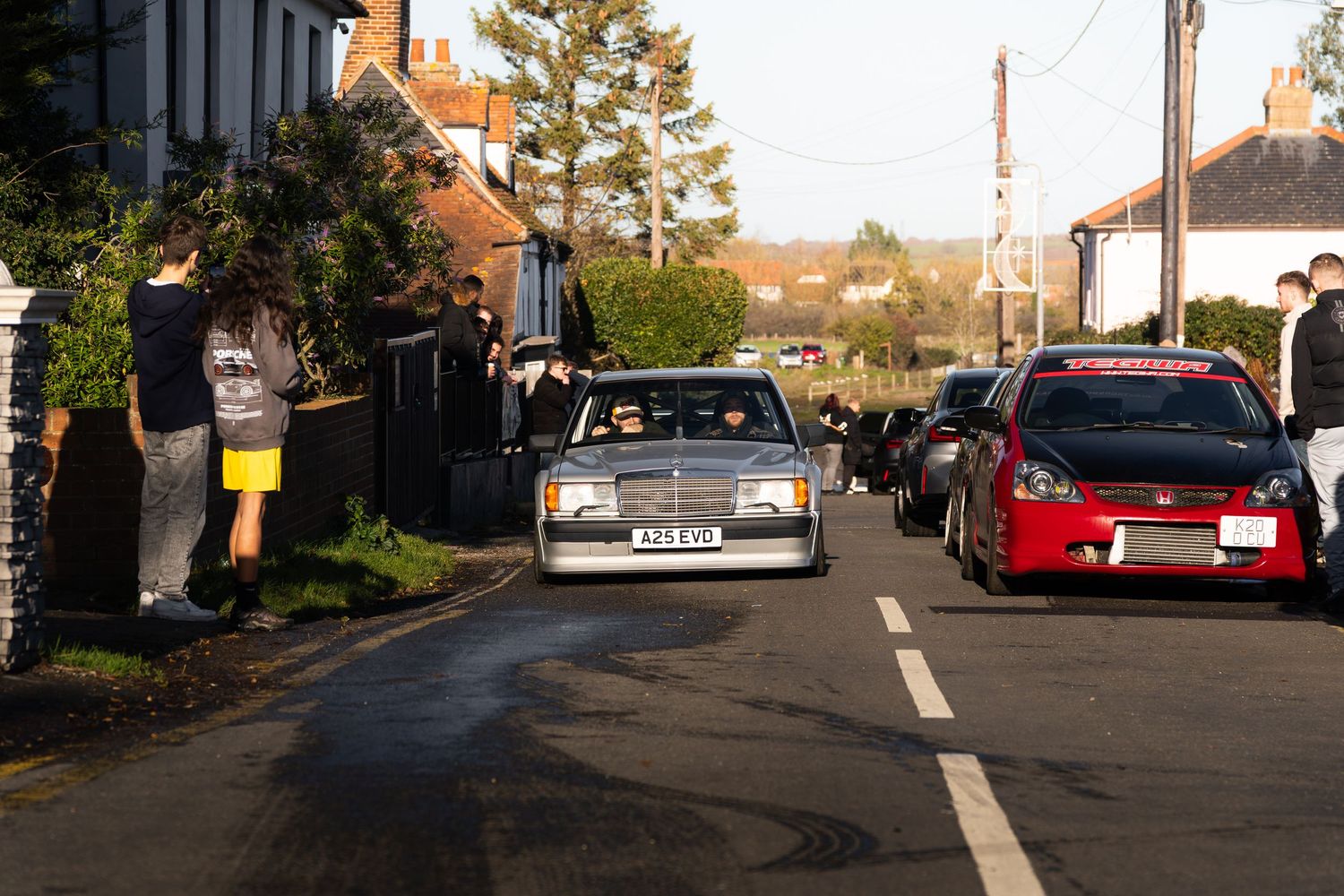 Classic white Mercedes 190E leading a convoy of cars through a village street.