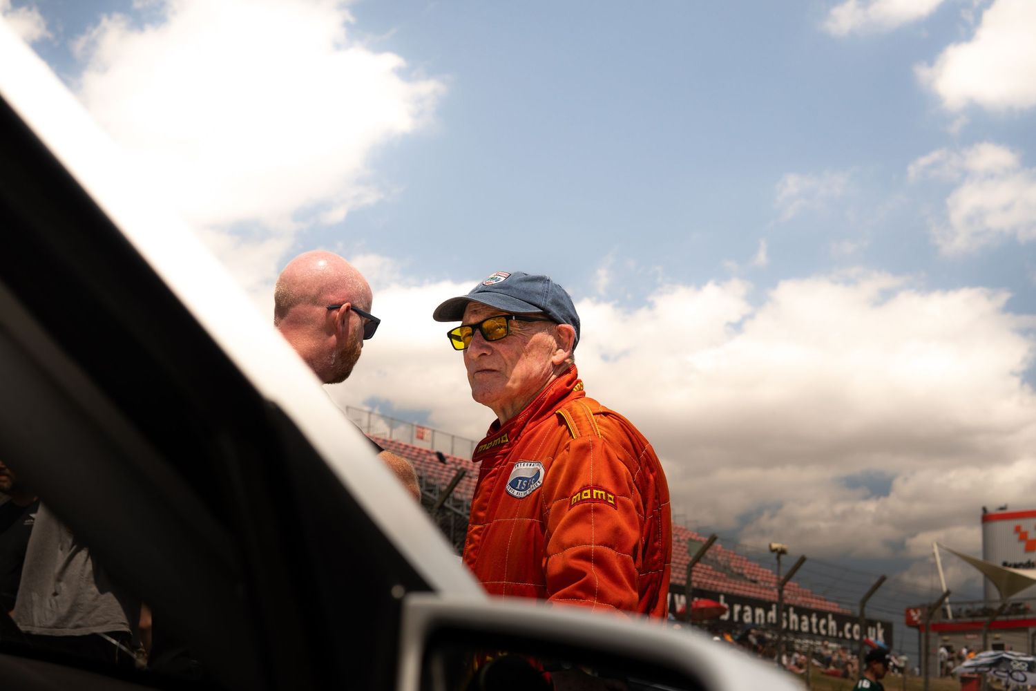 Racing driver in orange suit and blue cap at Brands Hatch circuit paddock.