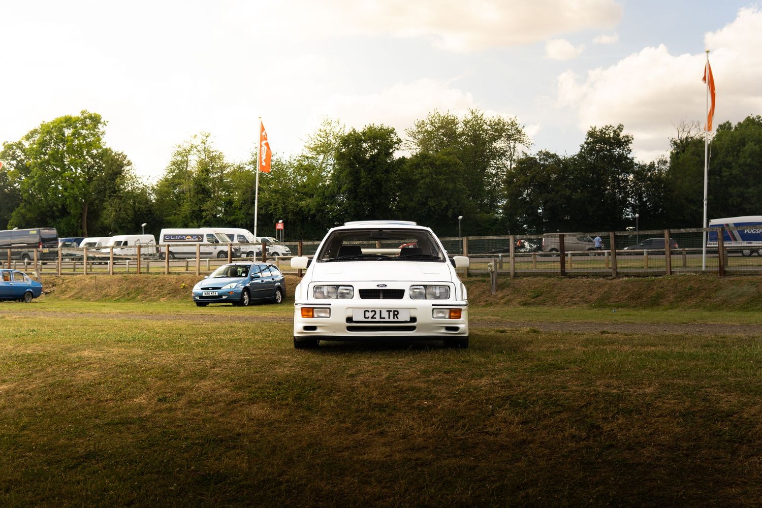 White Ford Sierra RS Cosworth parked on grass at a classic car show with flags and caravans in background.