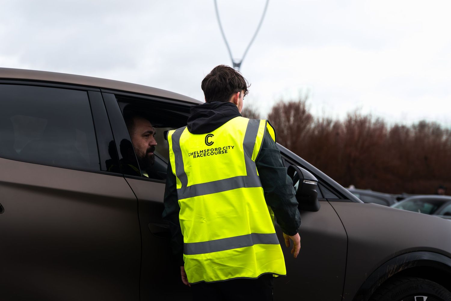 Security marshal in yellow hi-vis vest checking in a dark SUV at a car event.