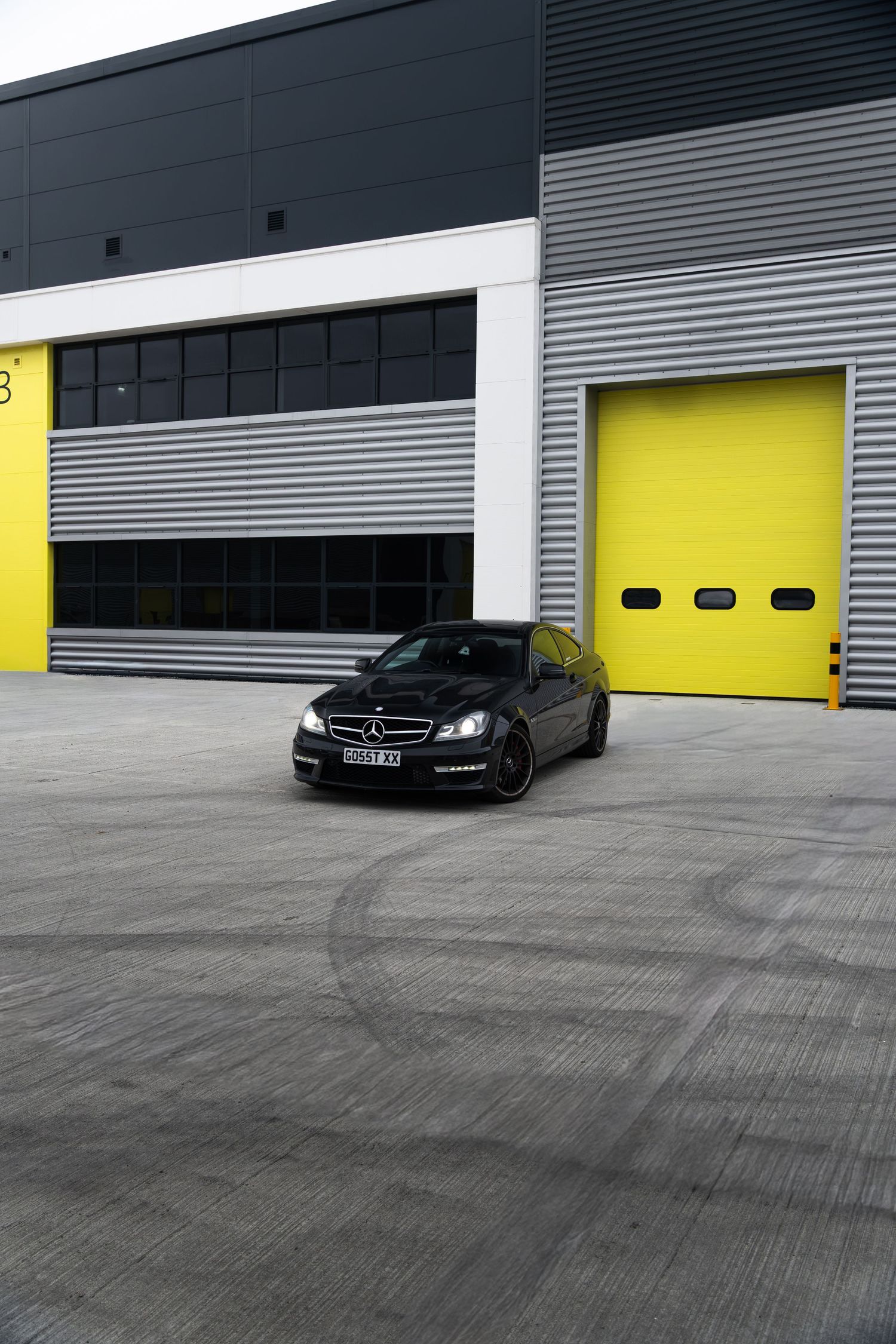 Black Mercedes-AMG C63 coupe parked in spacious concrete yard outside industrial building with yellow doors.