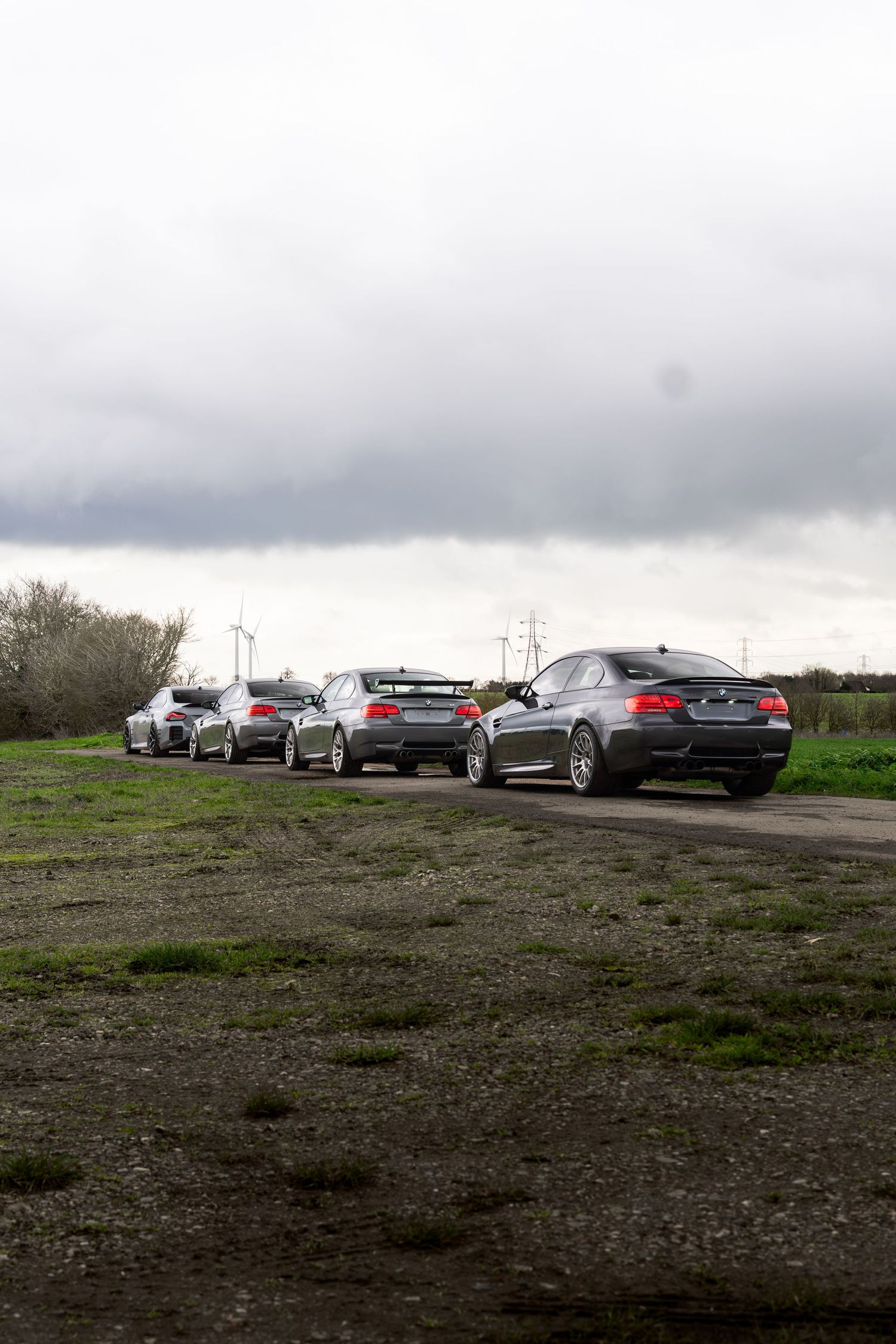 Four BMW M3 coupes lined up on a rural dirt road with green grass and wind turbines visible.