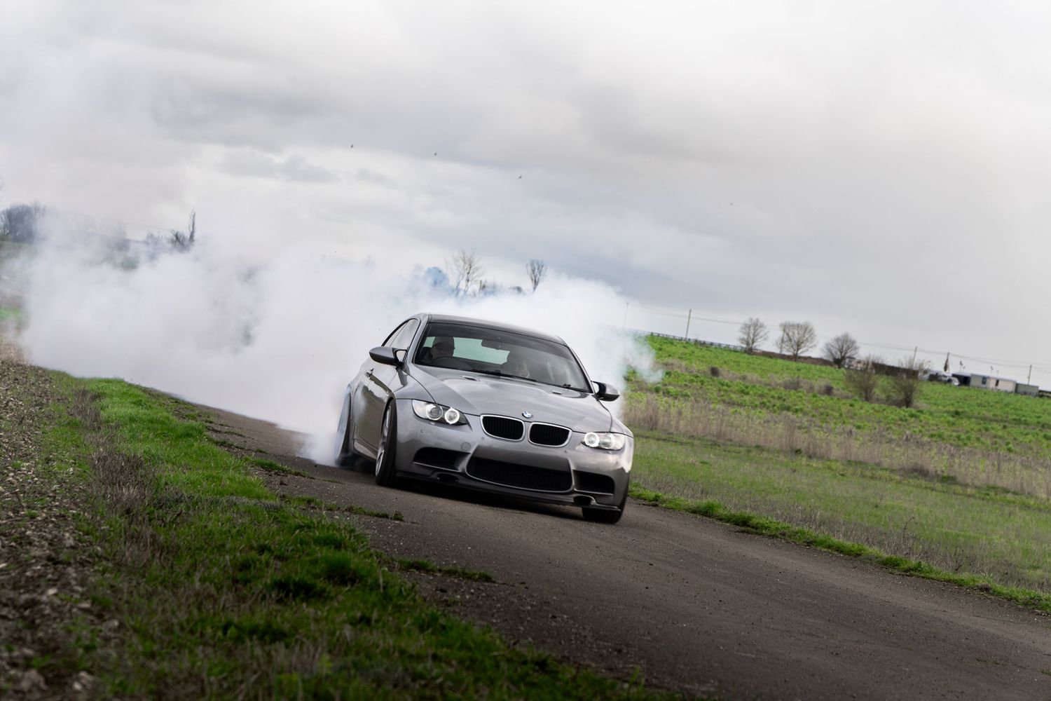 BMW M3 coupe performing a smoky drift on a country road with green fields behind.