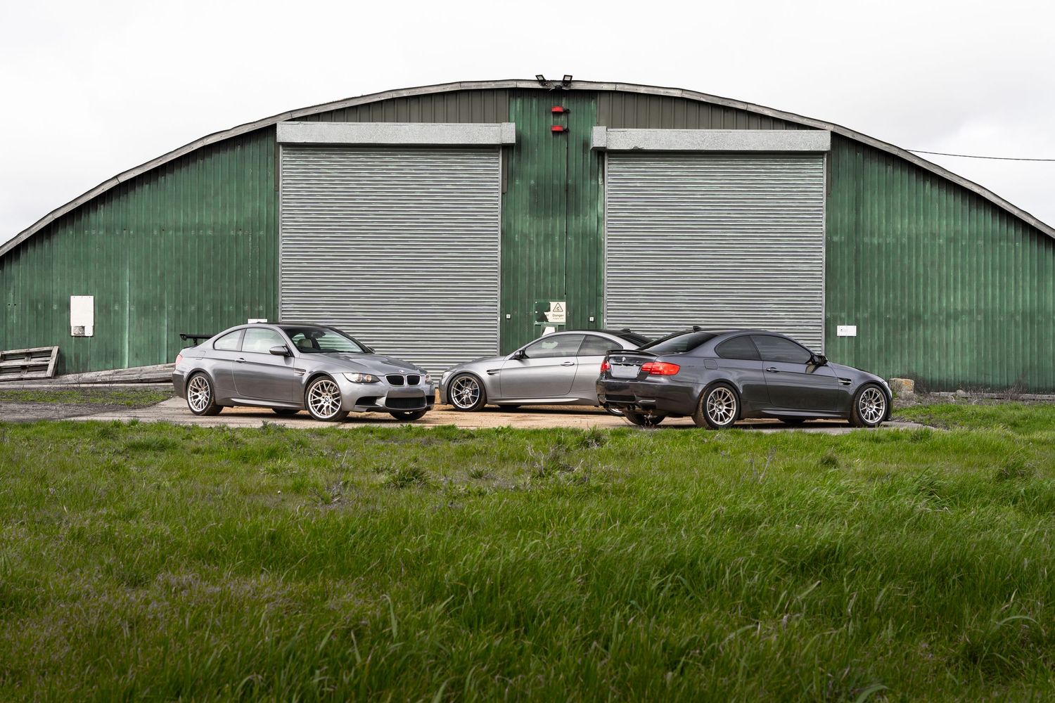 Three silver BMW M3 coupes displayed outside a green metal arched warehouse building.
