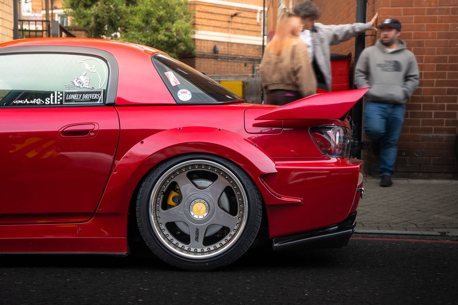 Red Mazda RX-7 with widebody kit and gold brake calipers on deep-dish alloy wheels.