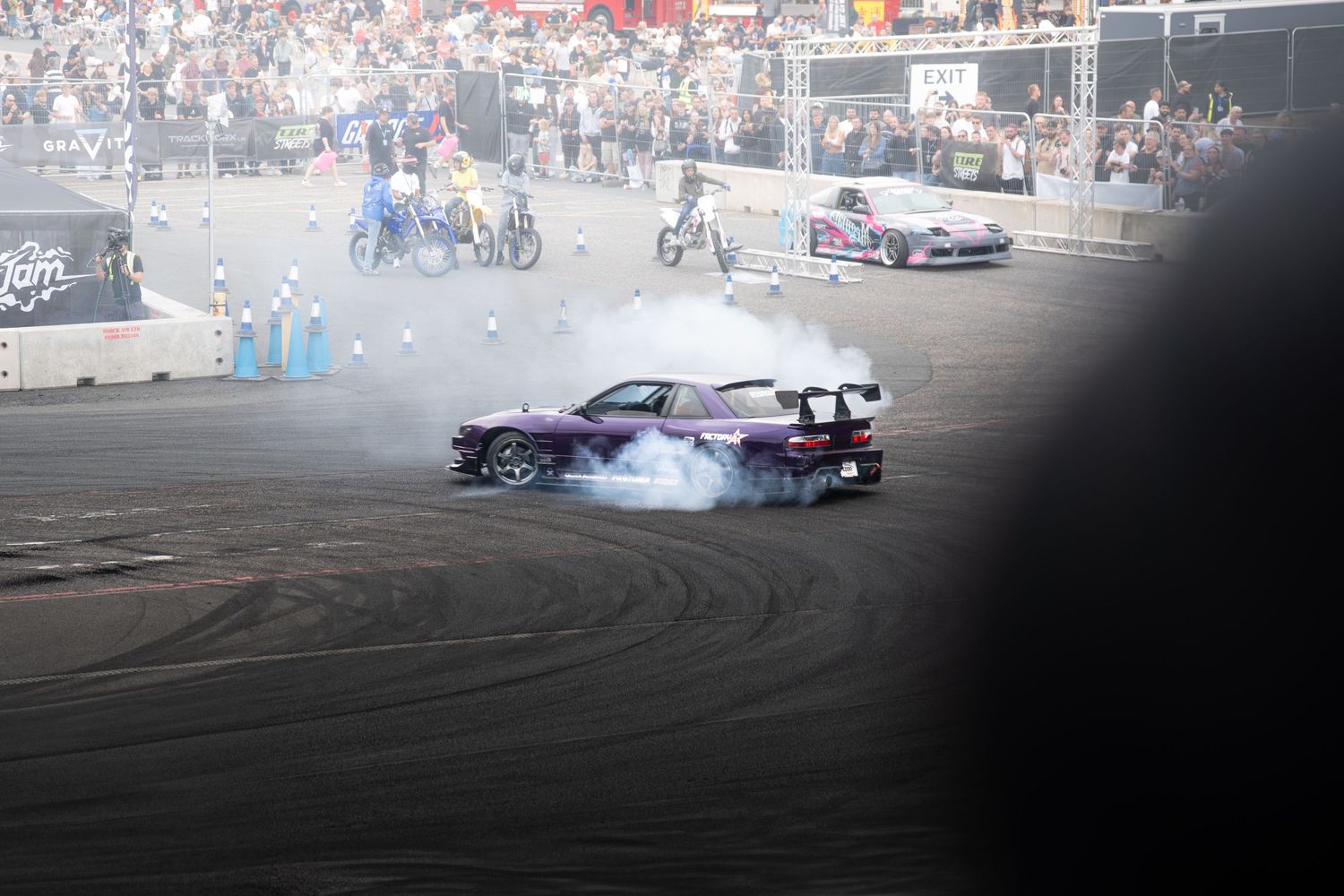 Purple drift car performing smoky burnout on a track in front of a large crowd.