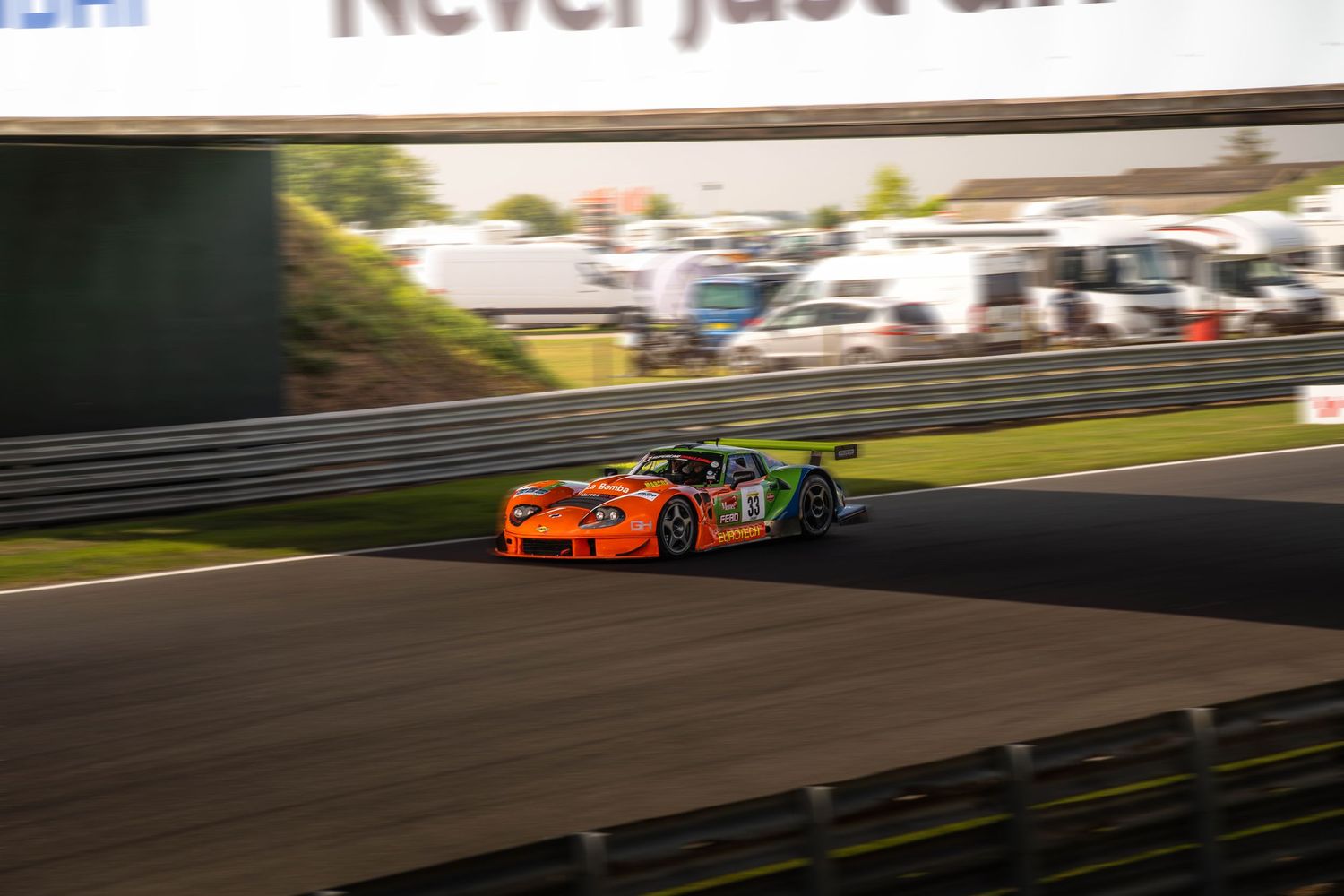 Orange GT race car speeding through a circuit under a bridge at dusk.