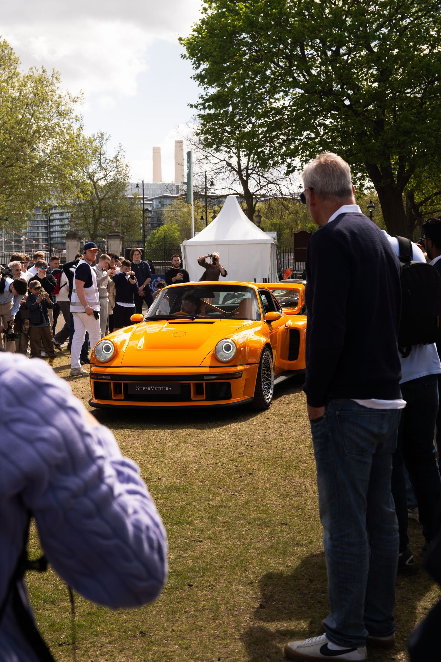 Orange RWB Porsche 930 Turbo on grass surrounded by photographers at outdoor car event.