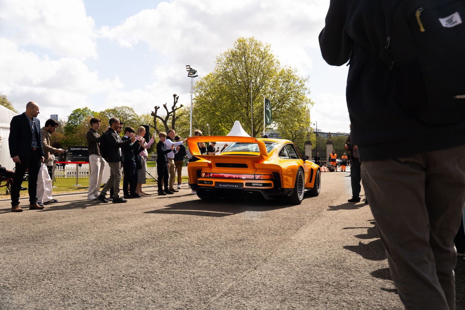 Orange wide-body RWB Porsche 930 Turbo driving past crowds at an outdoor car show.
