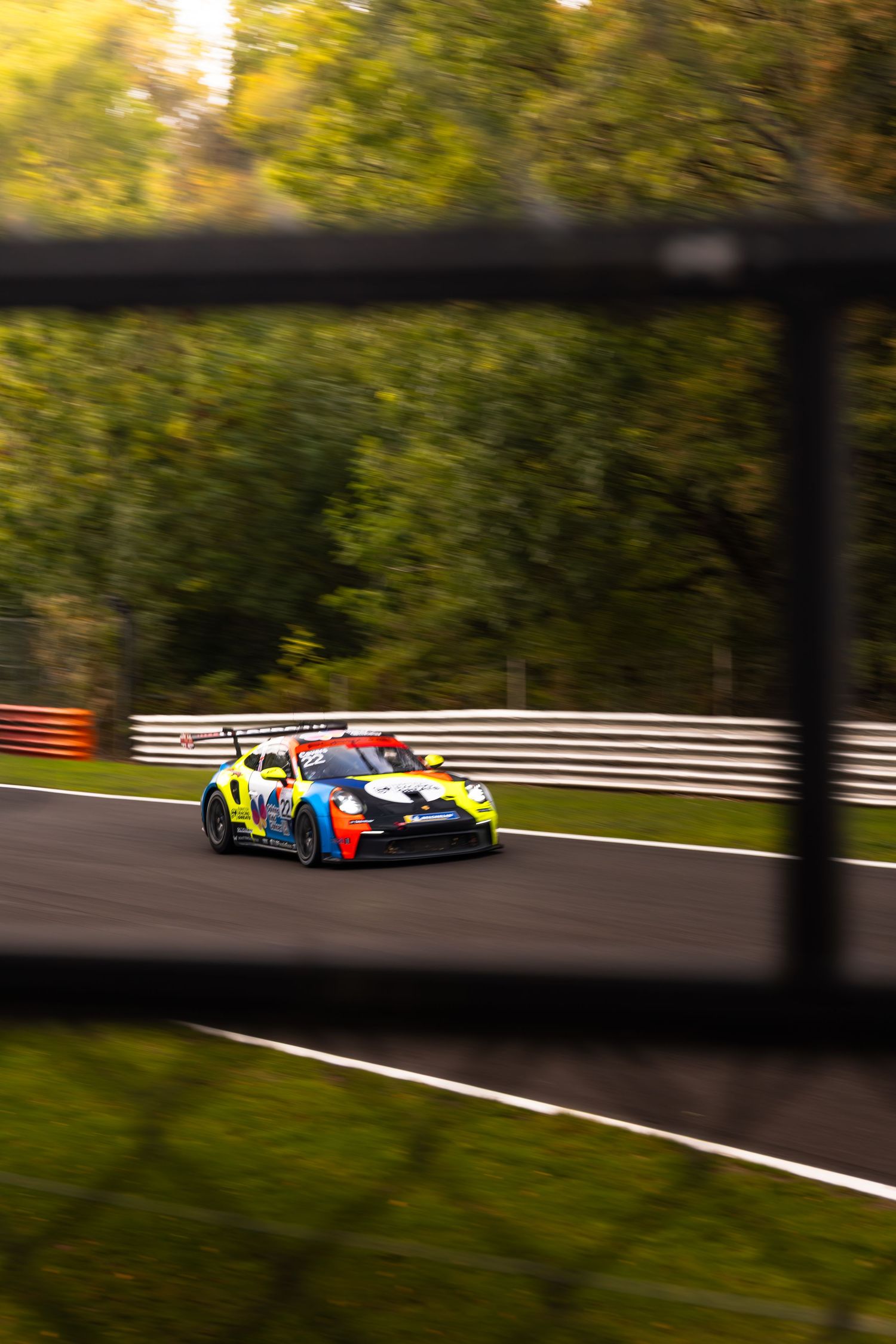 Colorful Porsche GT3 race car photographed through a fence on an autumn racing circuit.