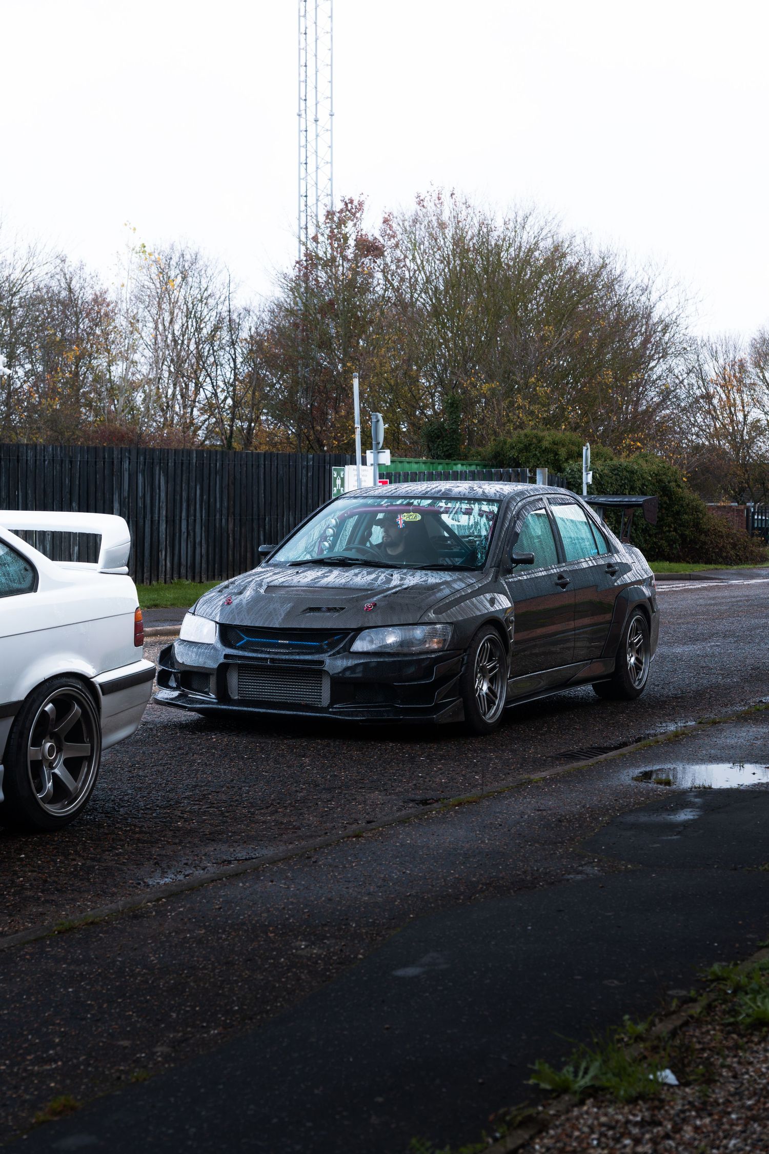 A dark modified Mitsubishi Lancer Evo parked on a wet road beside bare autumn trees.