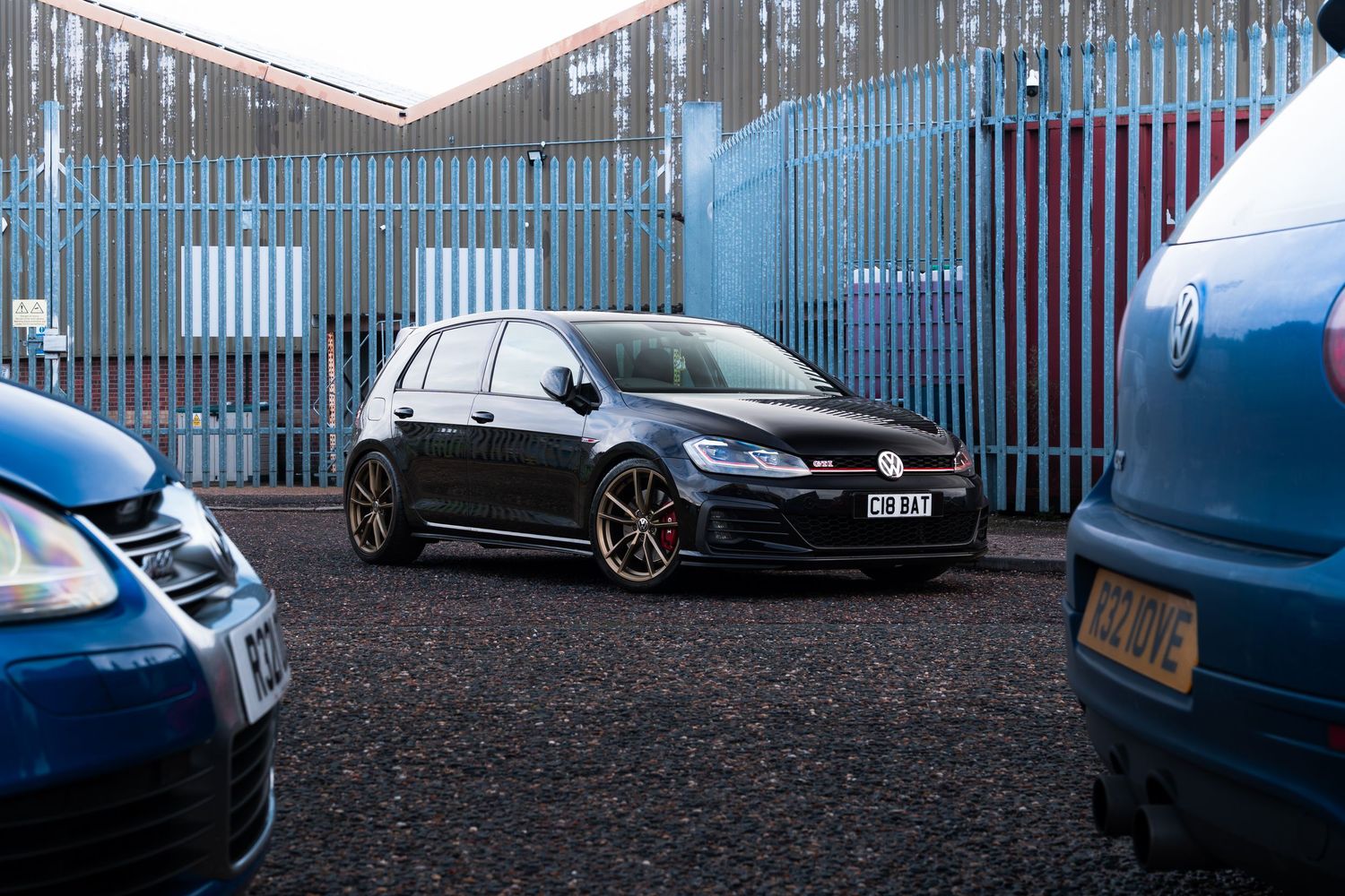 Black VW Golf GTI lowered on bronze alloy wheels parked in industrial yard between blue van and dark hatchback.