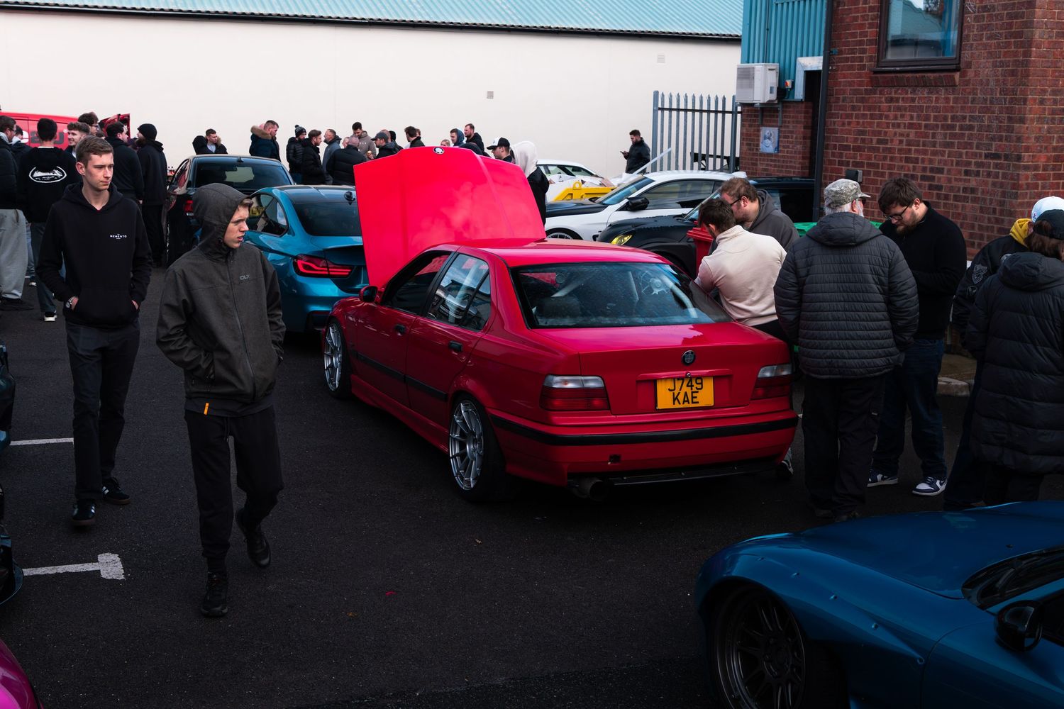 A red BMW E36 with hood open surrounded by car enthusiasts at an outdoor meet.
