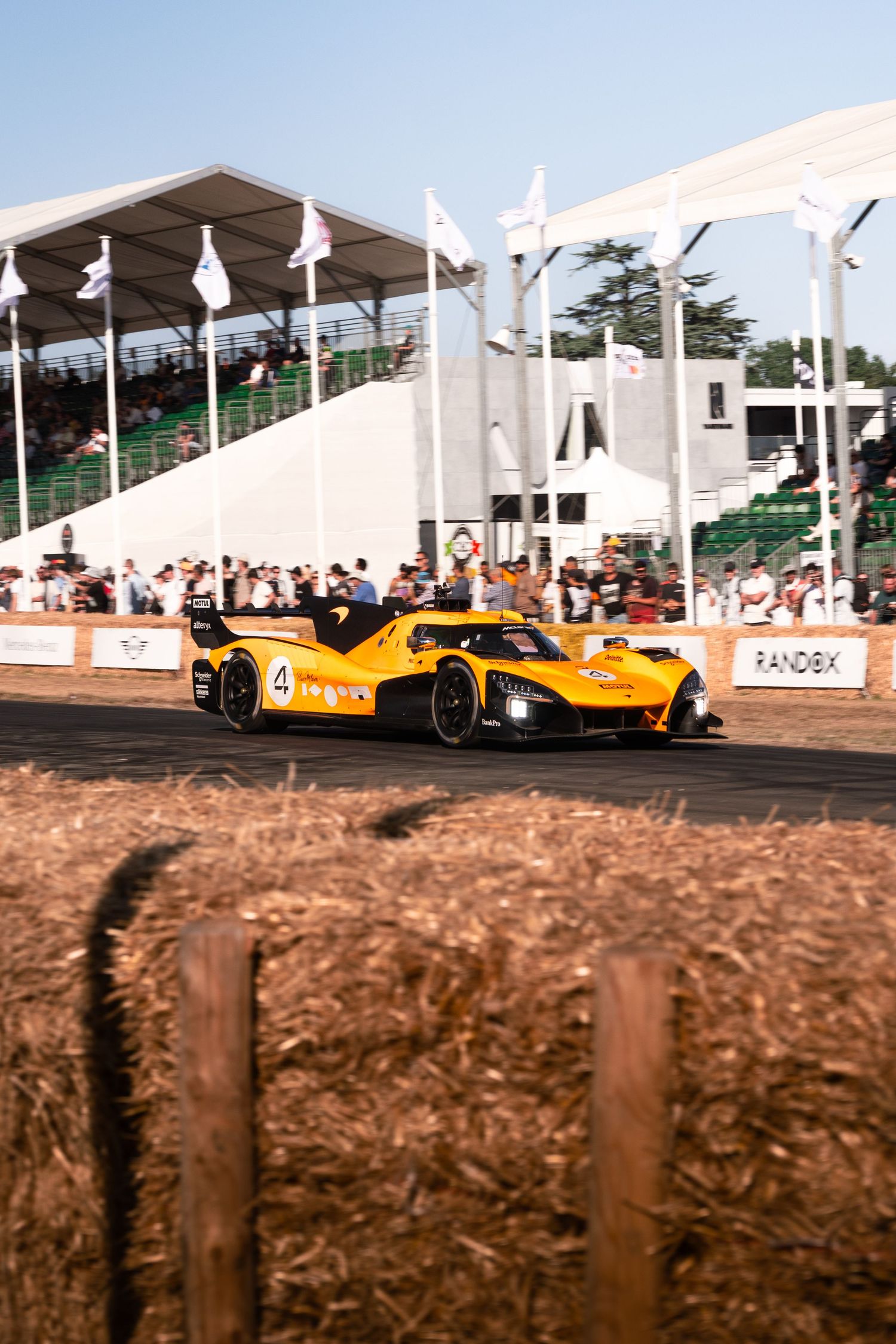 Yellow McLaren LMP racing car number 4 speeding past hay bales at Goodwood Festival of Speed with grandstands behind.