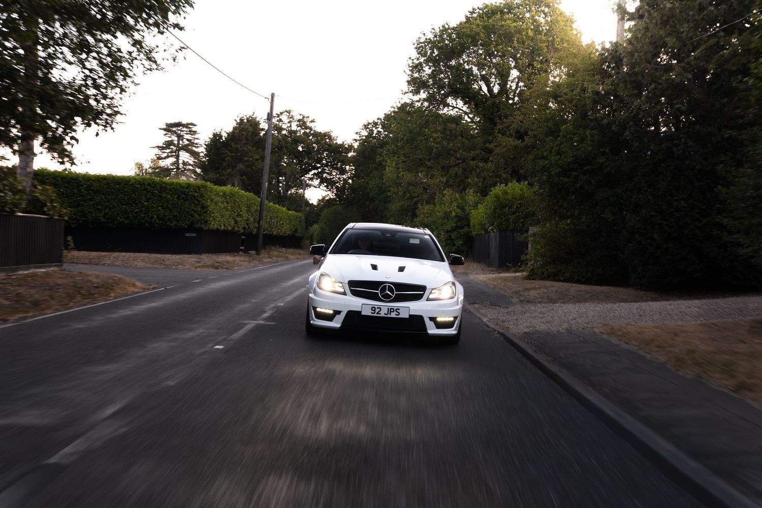 White Mercedes-Benz AMG speeding down a tree-lined road at dusk, captured from a front-facing perspective.