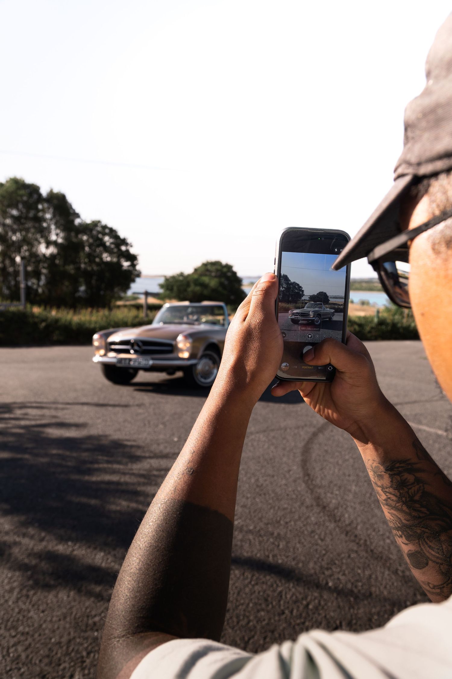 Tattooed person photographing a classic vintage car on smartphone in a parking lot with trees in background.