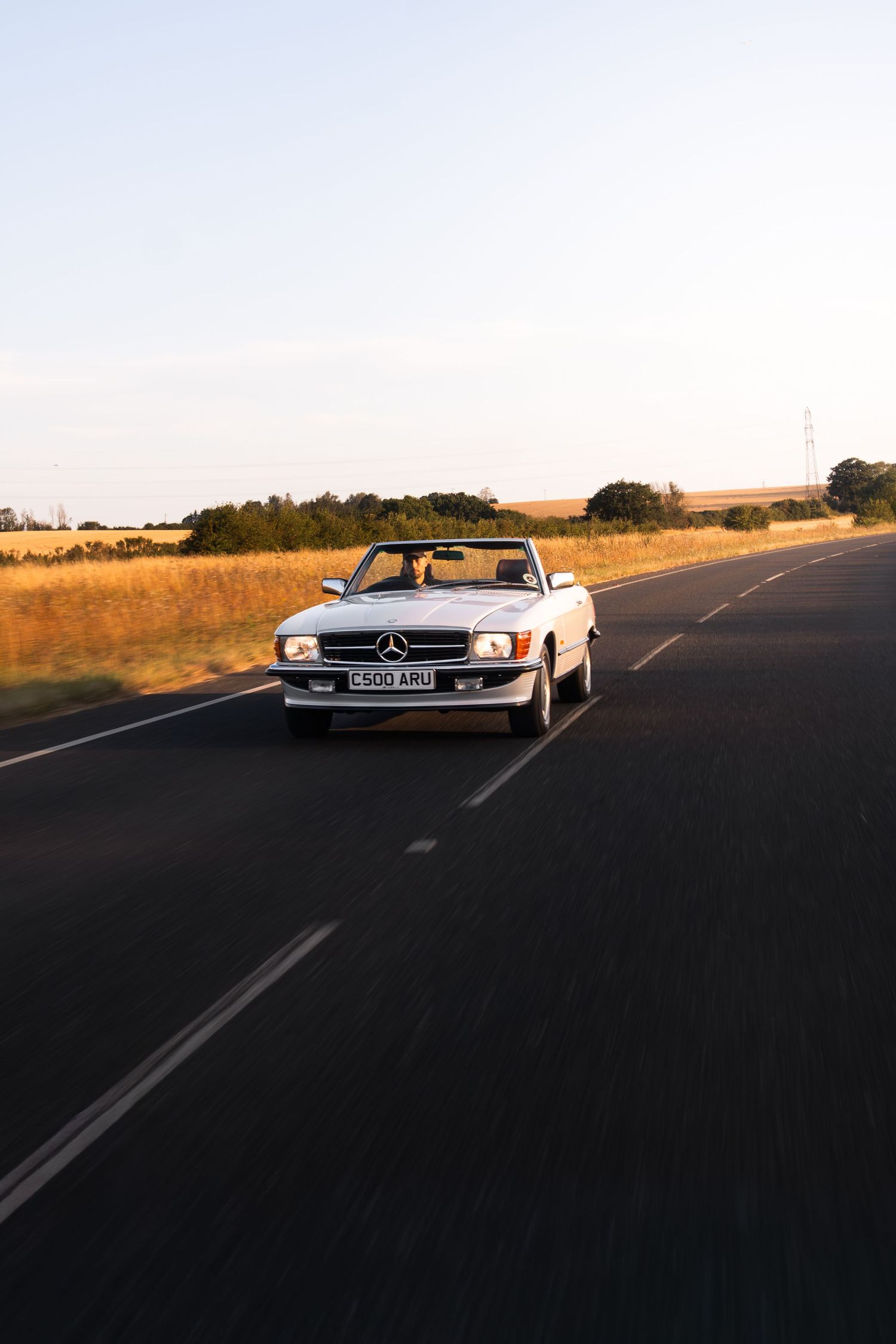 White Mercedes-Benz convertible classic car driving on open road through golden fields at sunset.