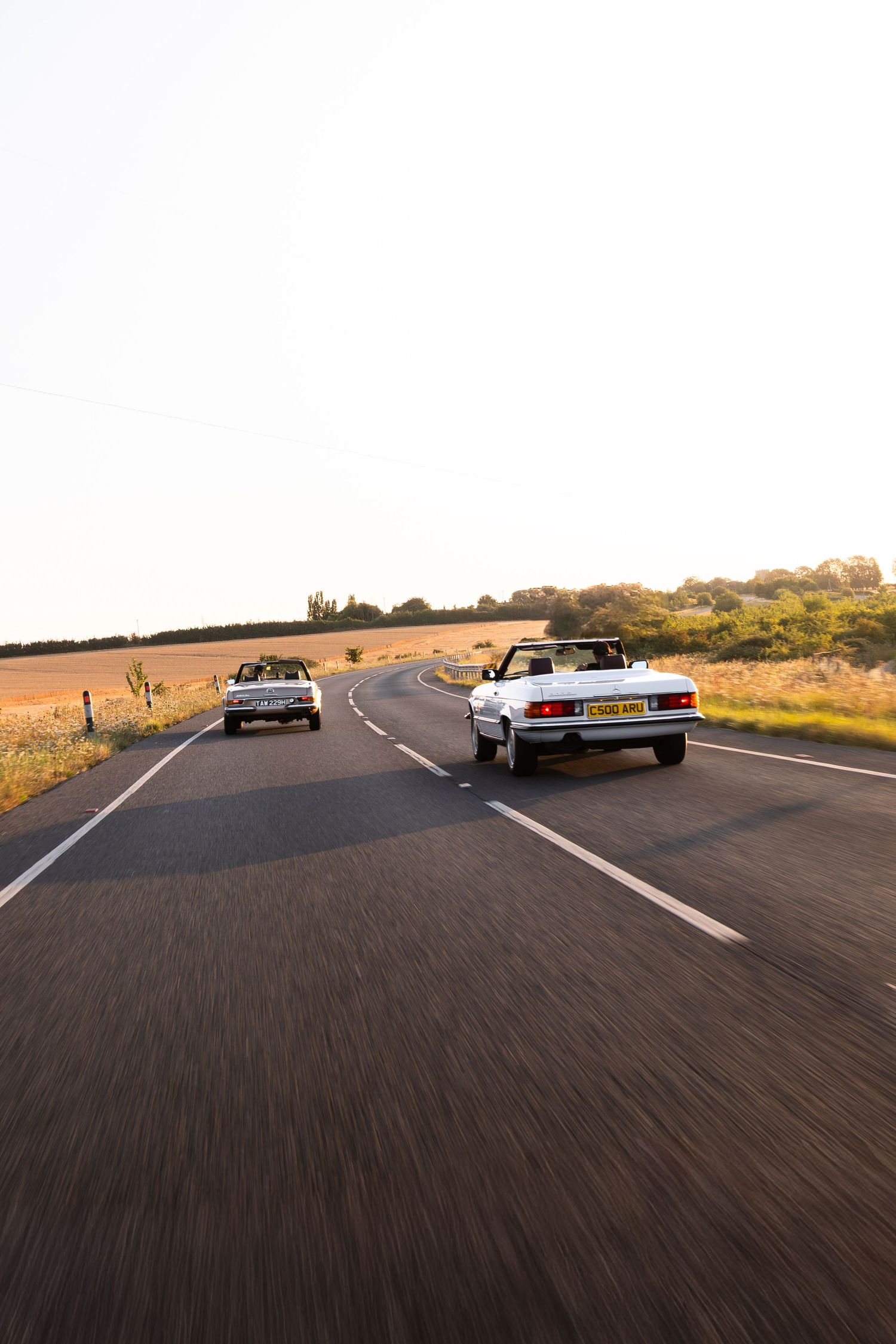 Two white convertible cars driving on a winding country road at golden hour sunset.