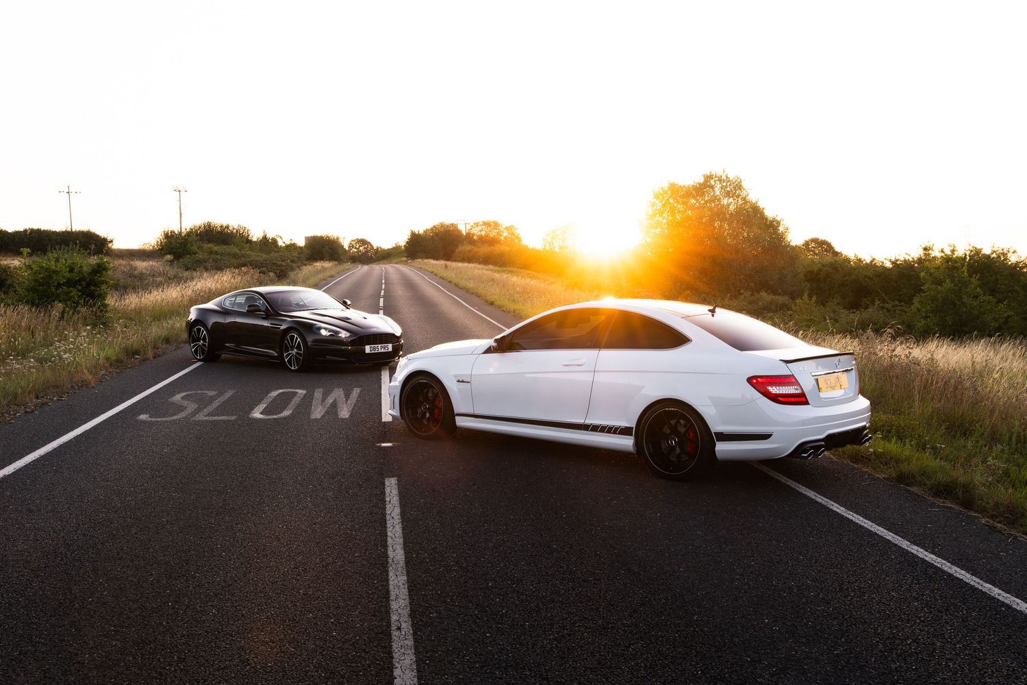 Two sports cars, white Mercedes C-Class and black coupe, parked on a rural road at golden sunset.