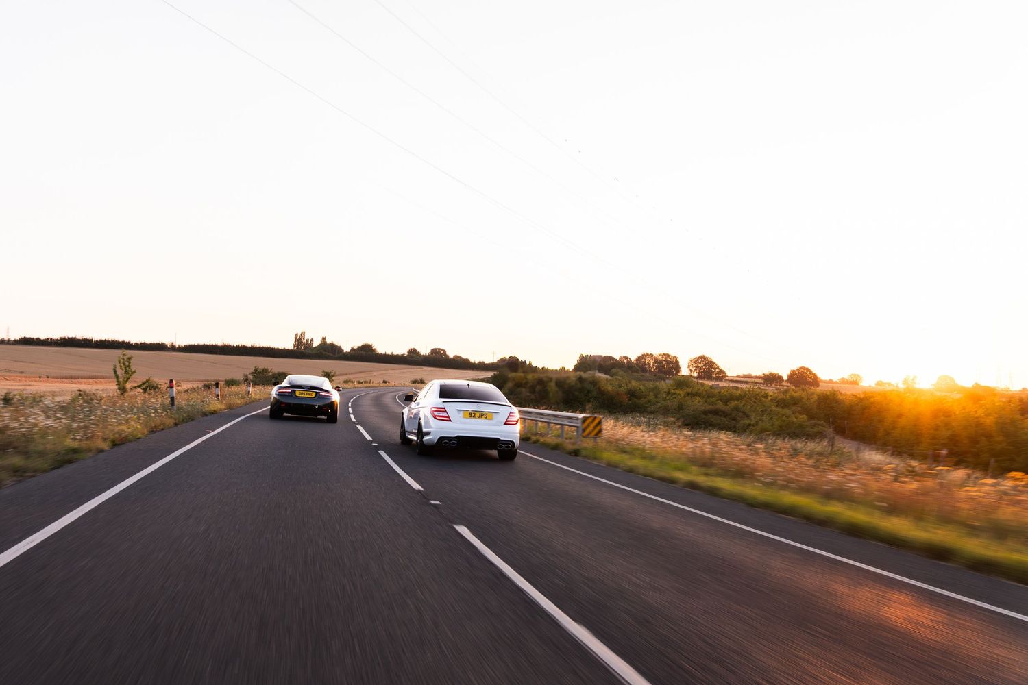 Two cars driving on a open road at sunset with golden fields on either side.