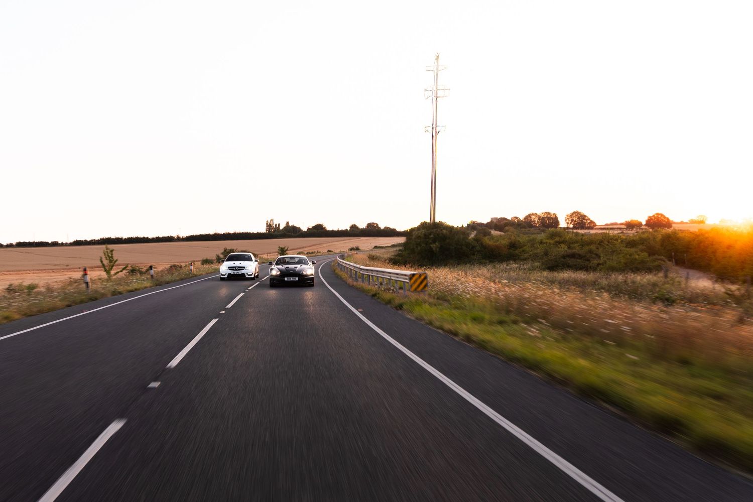 Two white cars driving on a straight rural road at sunset with golden light and power lines in the background.