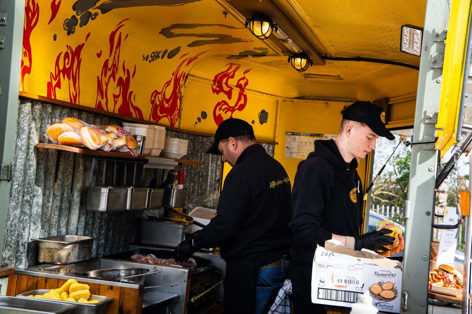 Two workers in black hoodies preparing food inside a colorful street food truck with flame decorations.