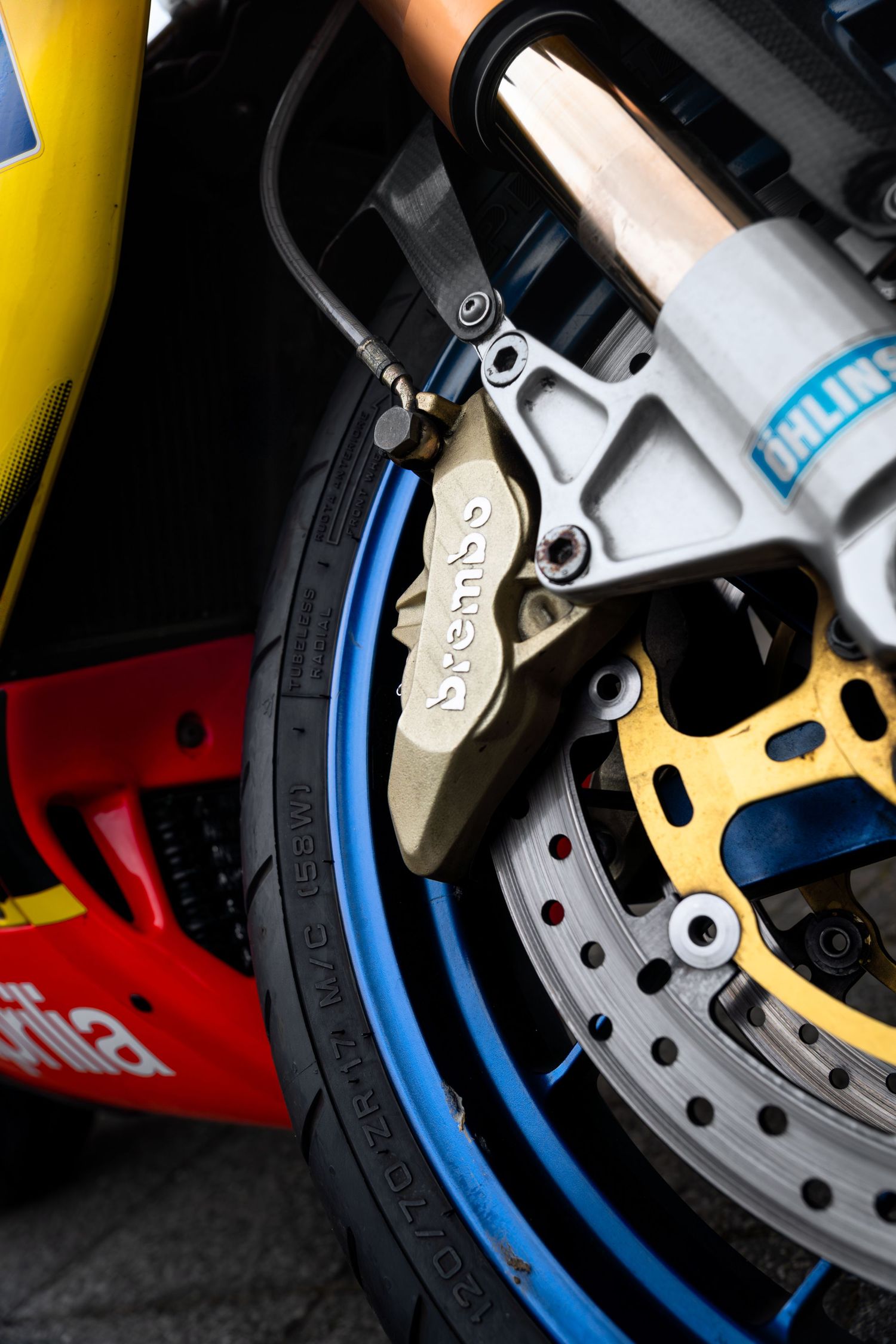 Close-up of Brembo brake caliper and gold rotor on blue-rimmed Aprilia motorcycle wheel.