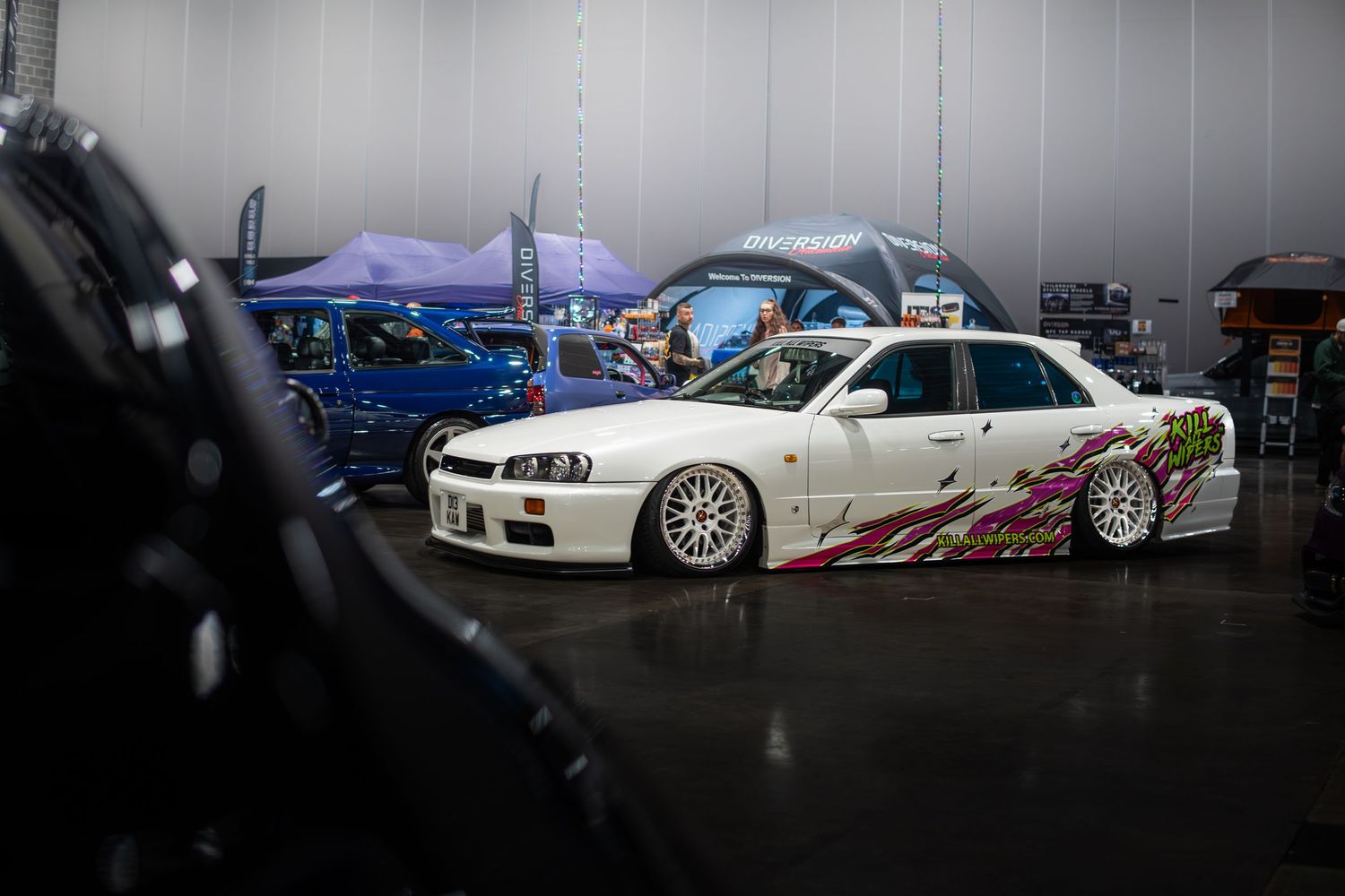 White Nissan Skyline R34 with vibrant graphics parked inside a car show venue.