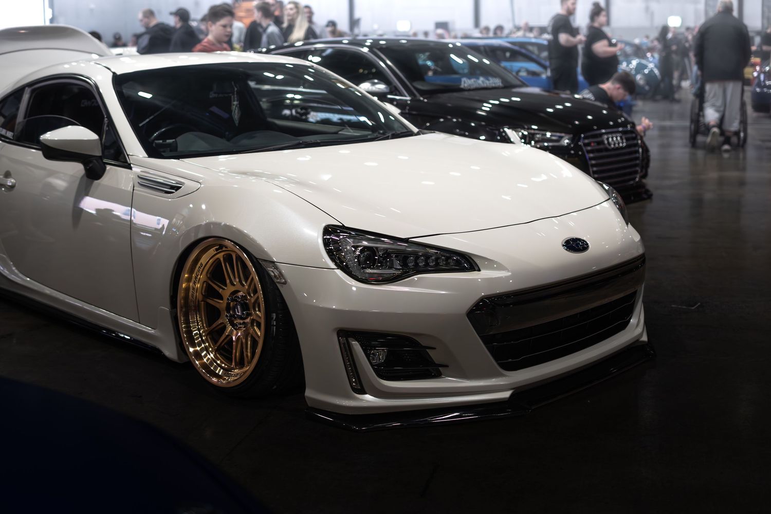 A white Subaru BRZ with gold BBS wheels and body kit on display at an indoor car show.