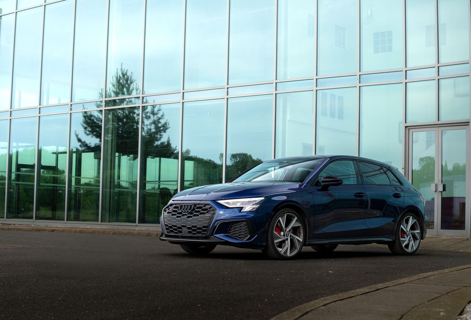 Navy blue Audi S3 Sportback parked beside a reflective glass building on a paved lot.